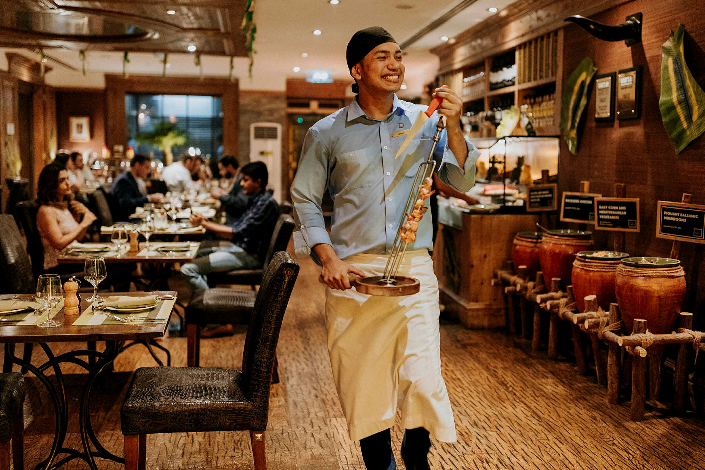 En tjener på en restaurant bærer en tallerken med grillede spyd. Andre gæster sidder ved borde i baggrunden. (AI-genereret tekst)