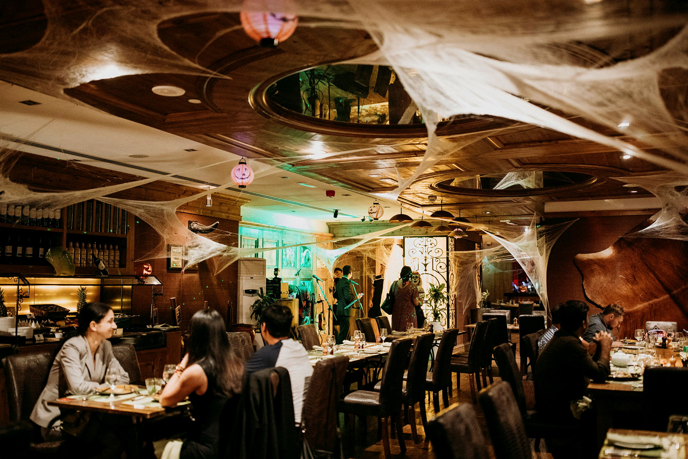 En restaurantinteriør dekoreret med spindelvævslignende pynt og belysning, med gæster ved borde og personale i baggrunden. (AI-genereret tekst)