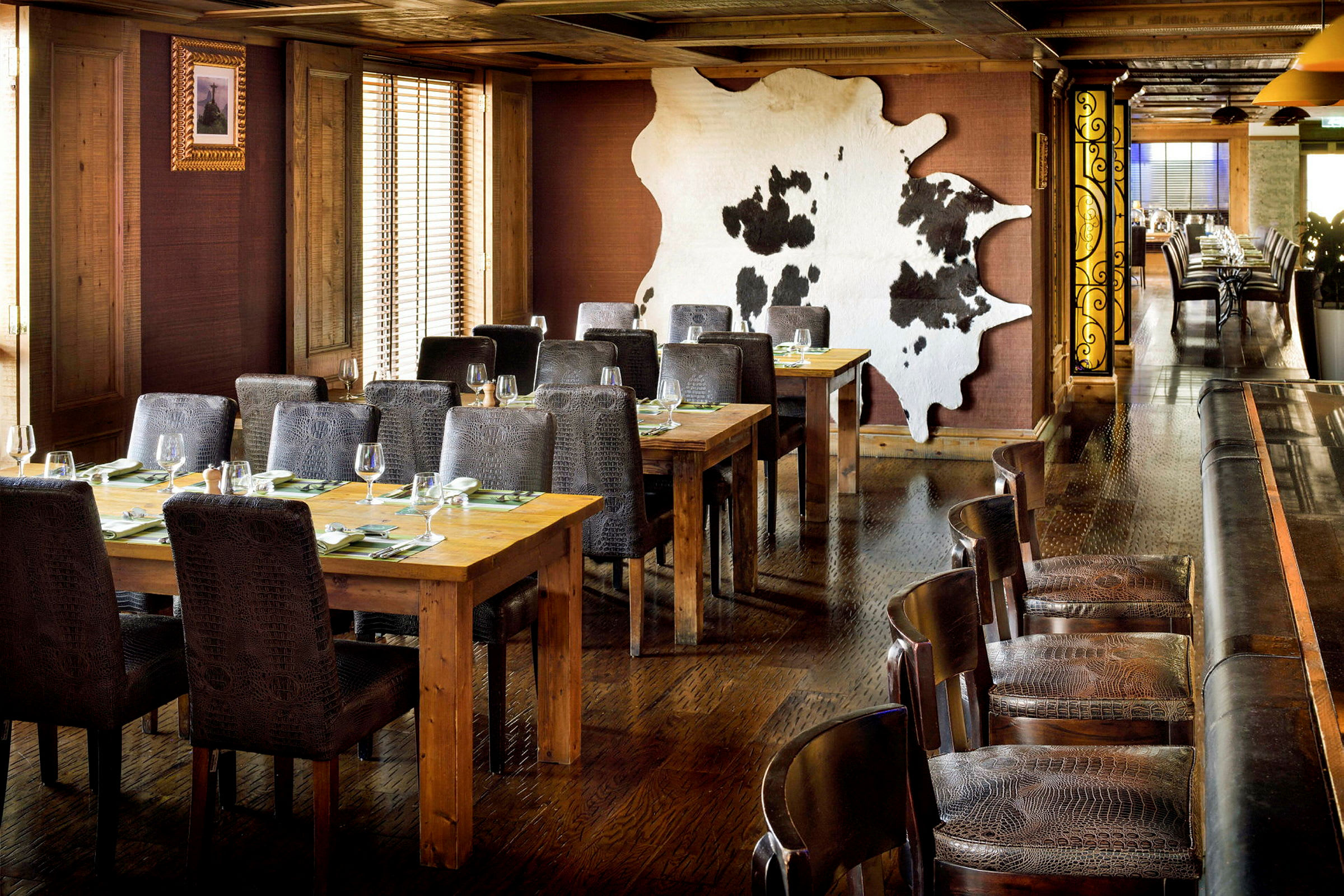Et restaurantområde med træborde og polstrede stole. En vægdekoration i form af en dyrehud ses i baggrunden. (AI-genereret tekst)