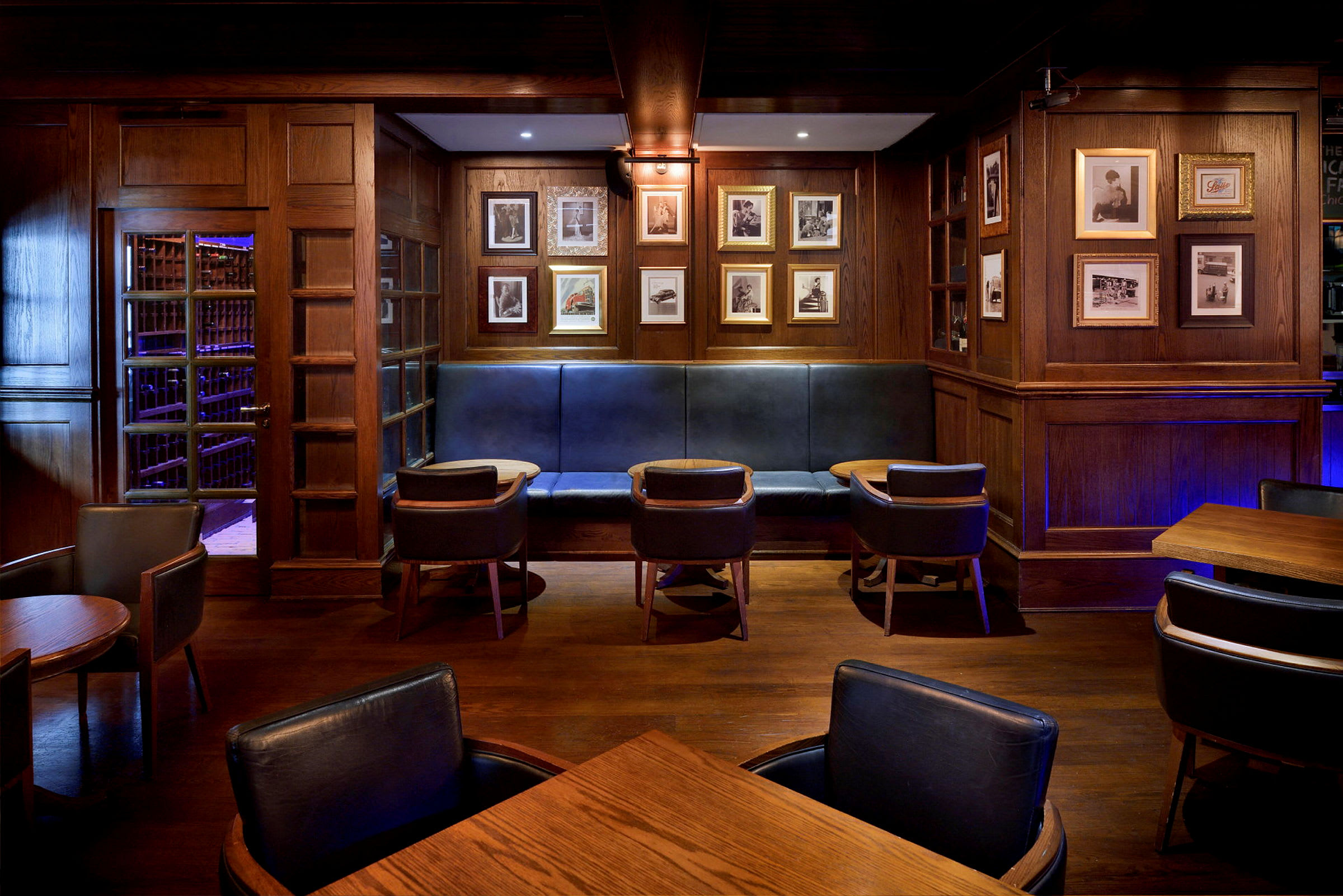 Et værelse indrettet som en lounge eller bar med træpaneler, indrammede billeder på væggen, polstrede bænke og stole samt små borde. (AI-genereret tekst)