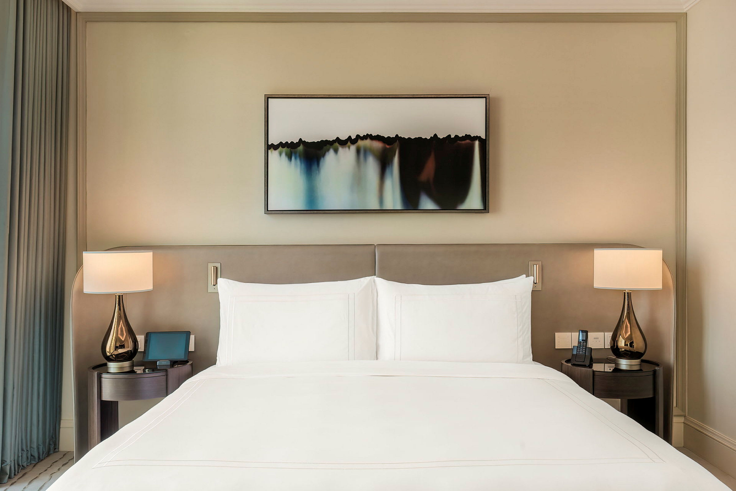 En velredt hotelseng med to natborde og lamper på hver side, samt et billede over sengens hovedgærde. (AI-genereret tekst)