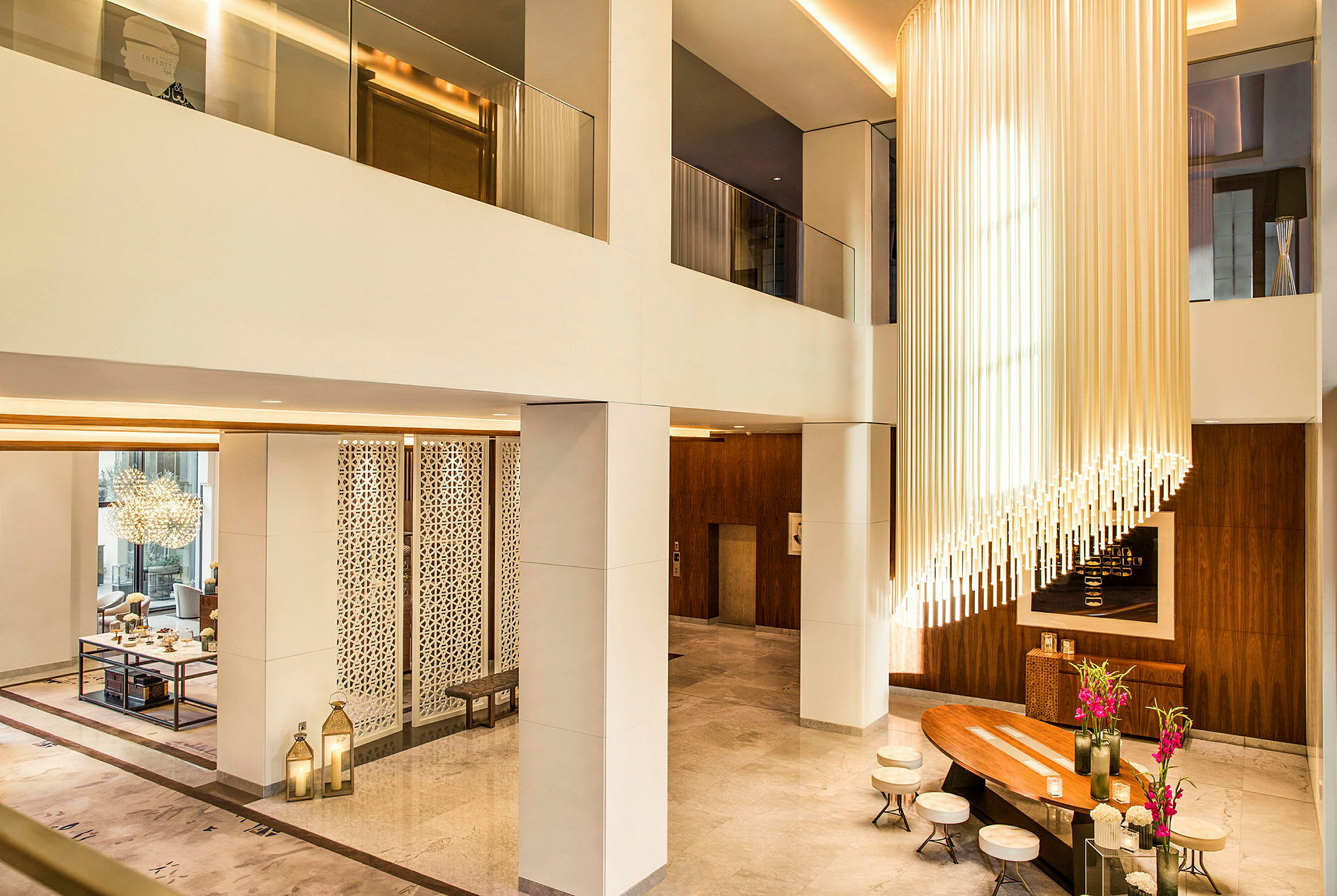 En elegant hotellobby med højt til loftet, moderne indretning og en lysekrone af lodrette rør. Der er siddepladser omkring et træbord med blomster. (AI-genereret tekst)
