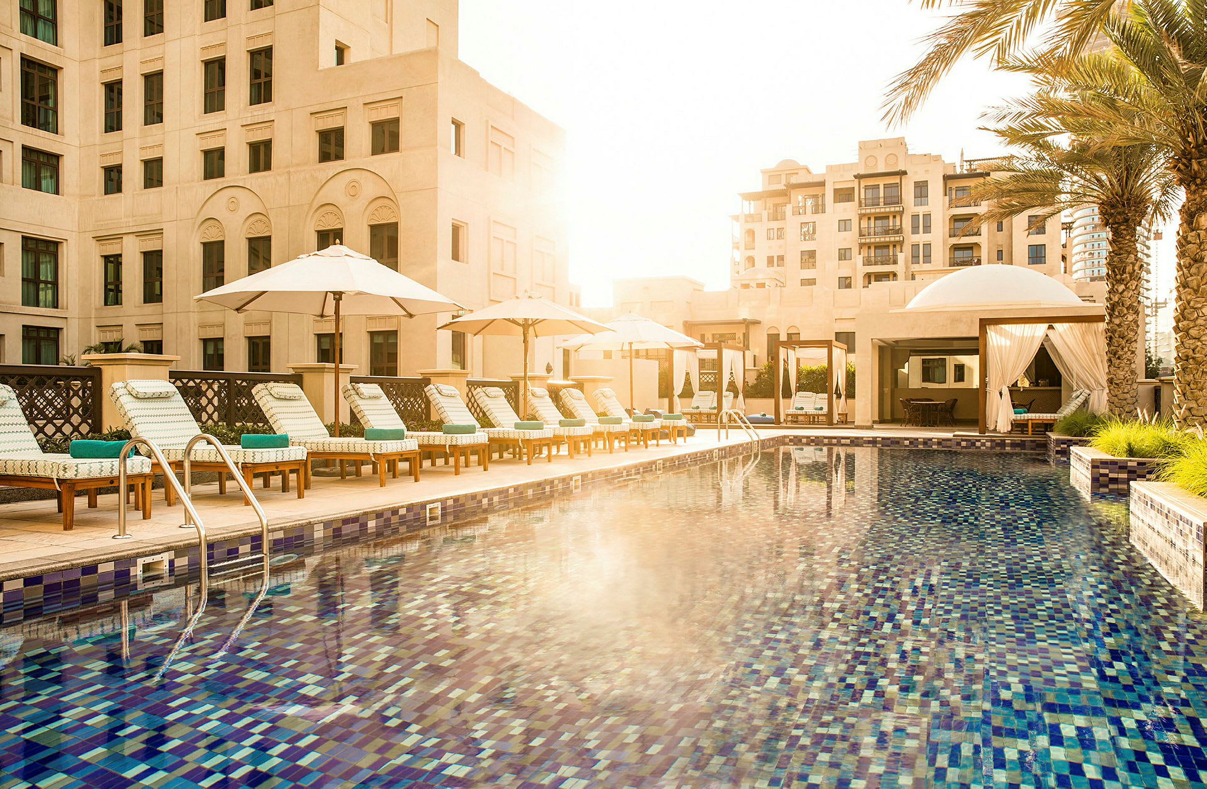 En hotelpool med liggestole og parasoller, omgivet af bygninger og palmer. (AI-genereret tekst)