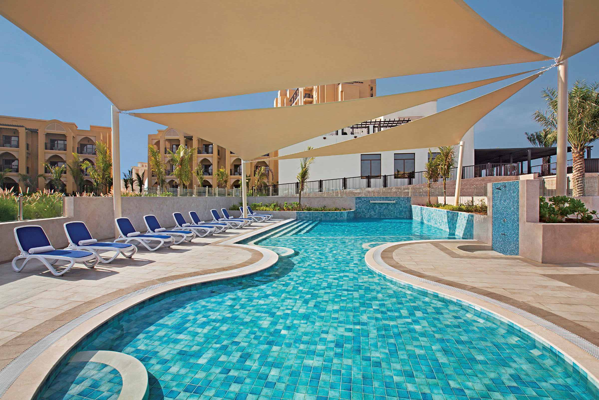 En pool med blåt vand, lyse beige liggestole og let skygget stof ovenover. I baggrunden ses bygninger. (AI-genereret tekst)