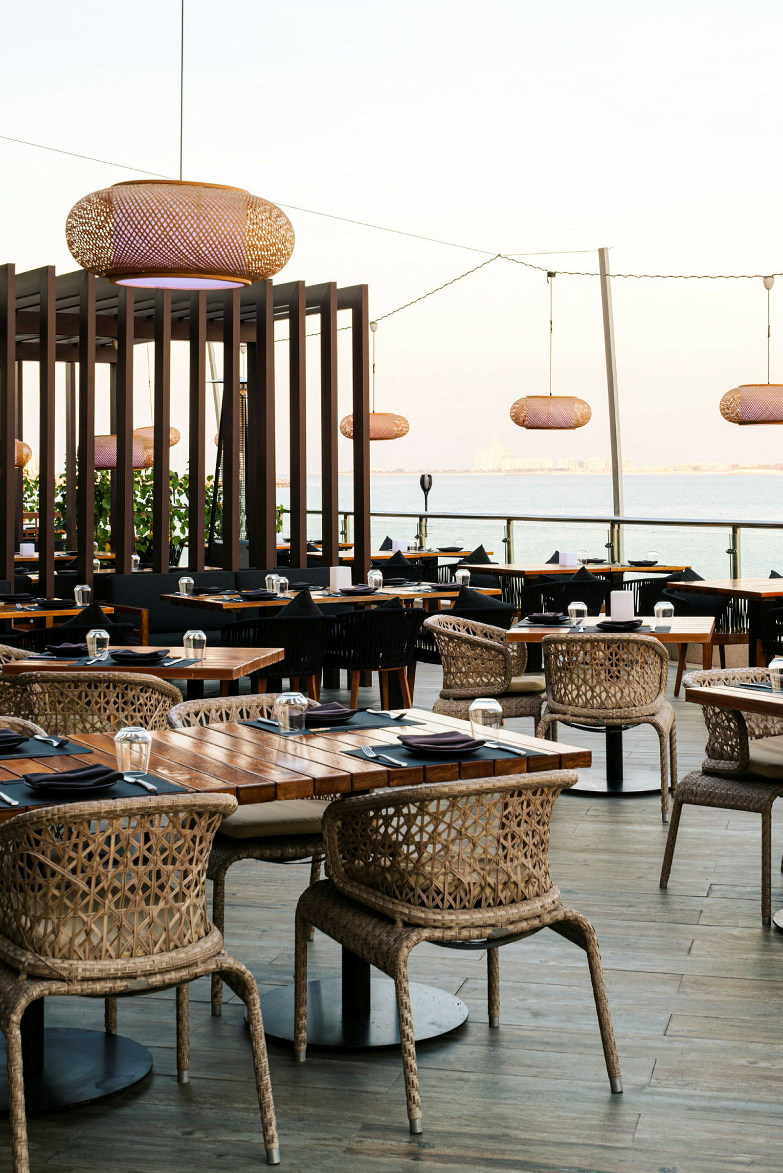 En udendørsrestaurant med træmøbler, rottingstole og borde klargjort med sorte servietter og glas. Overfor havet. (AI-genereret tekst)