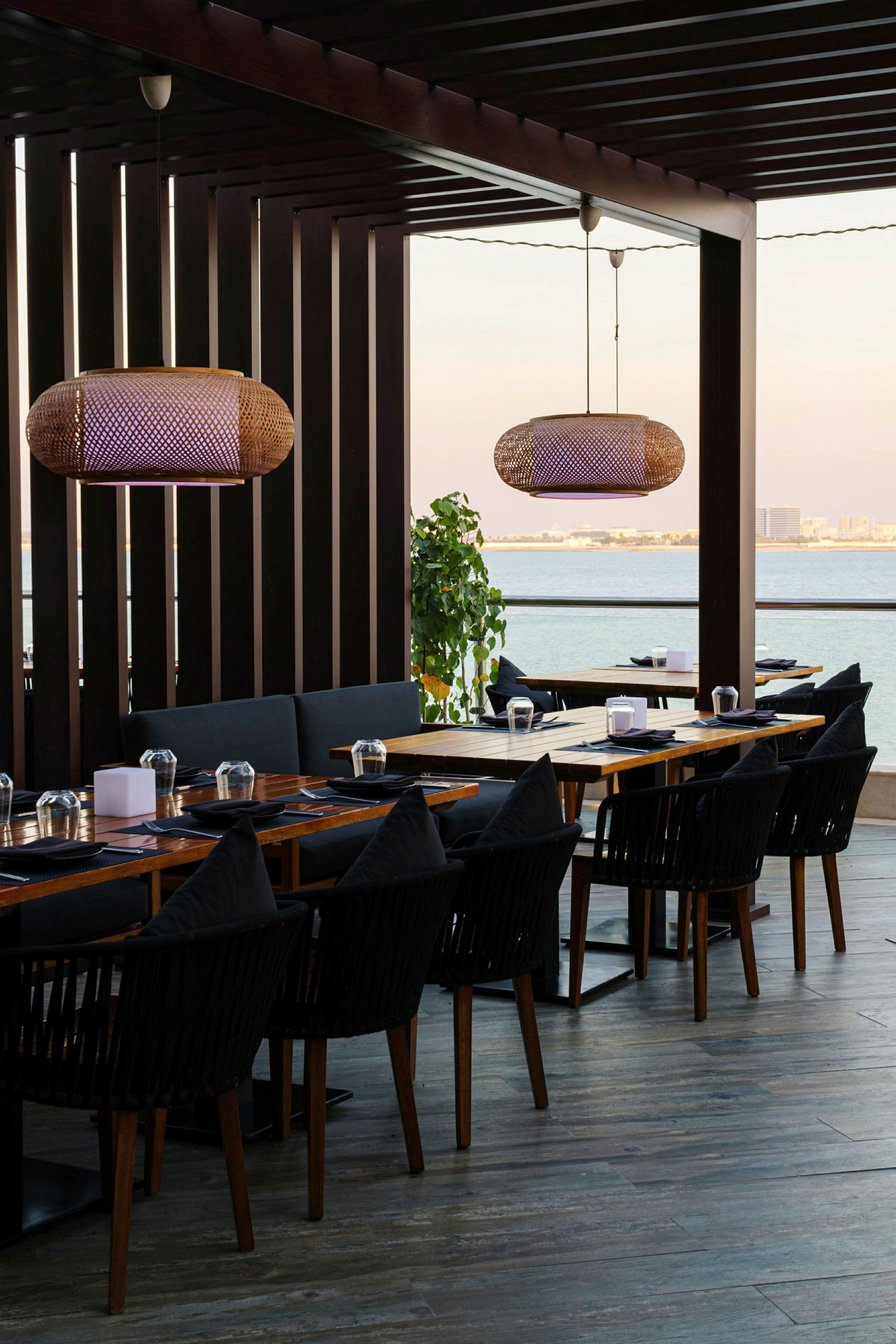 Udendørs serveringsområde med bord med træplader og sorte stole, omgivet af skygger og udsigt over havet. (AI-genereret tekst)
