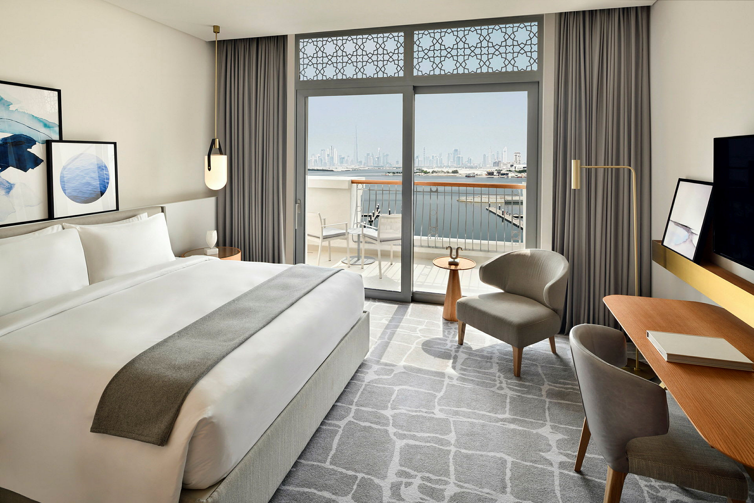 Et moderne indrettet hotelværelse med en stor dobbeltseng og vinduer, der åbner ud til en balkon med udsigt over en byens skyline og vand. (AI-genereret tekst)