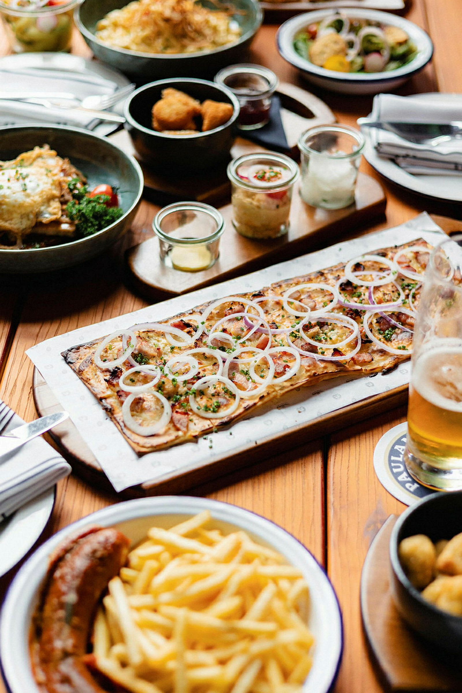 Et bord dækket med forskellige retter, herunder en pizza med løg, pommes frites og en pølse. (AI-genereret tekst)