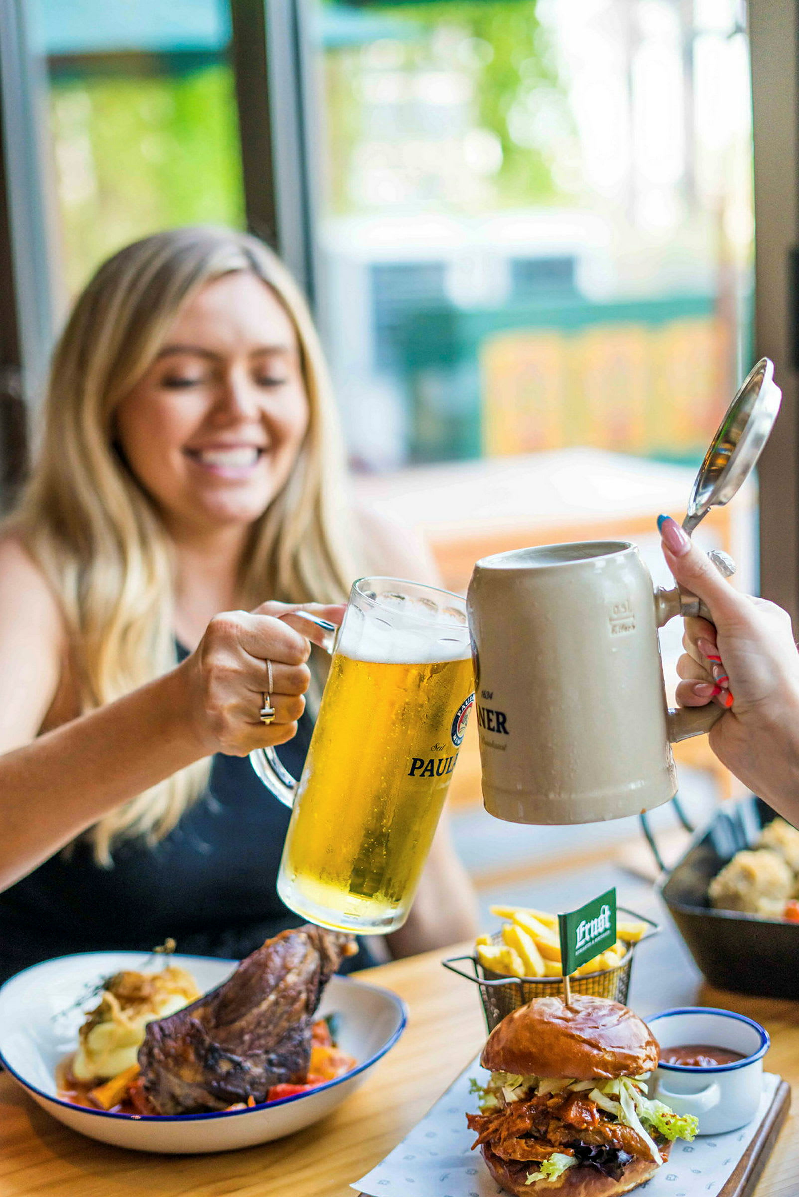 To fyldte ølglas skåles, og på bordet er tallerkener med mad inklusive burger, pommes frites og kød. (AI-genereret tekst)