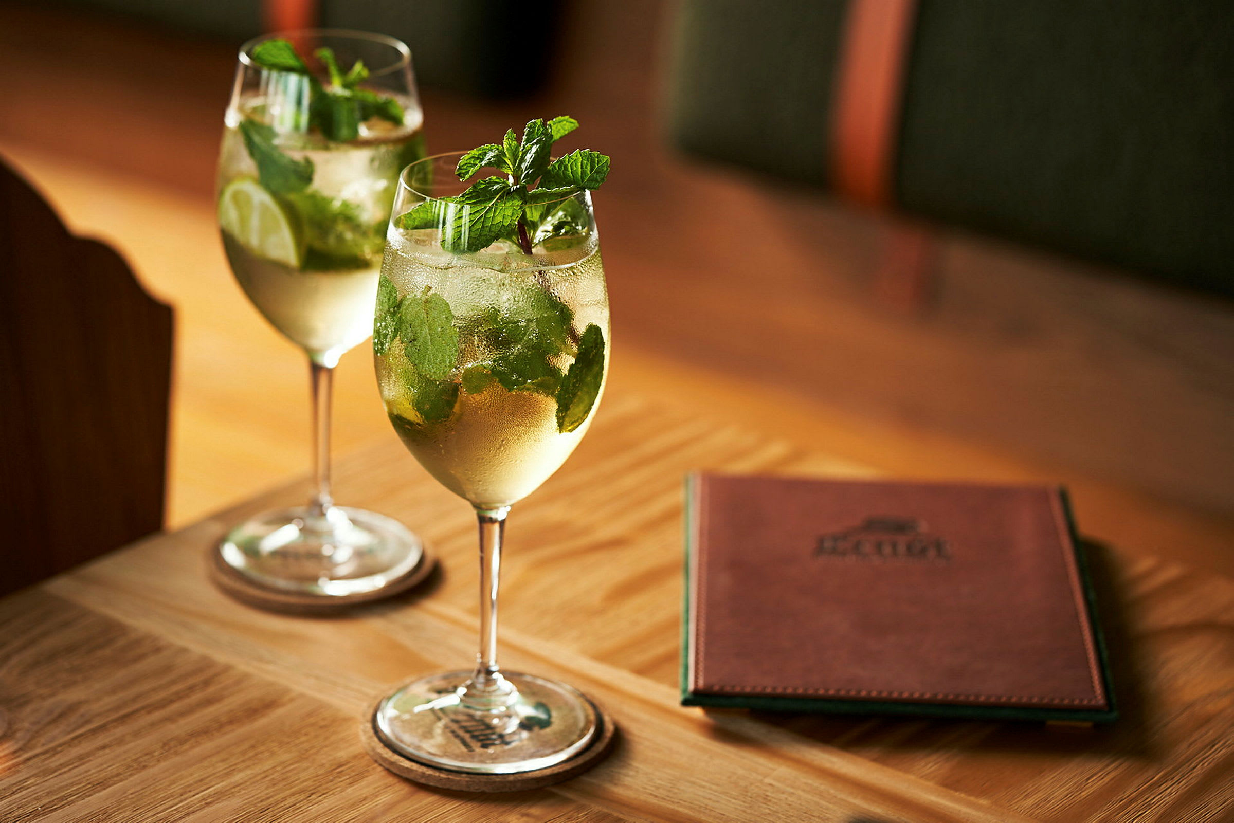 To glas med drinks indeholdende mynte og lime står på et bord ved siden af en menuhæfte. (AI-genereret tekst)