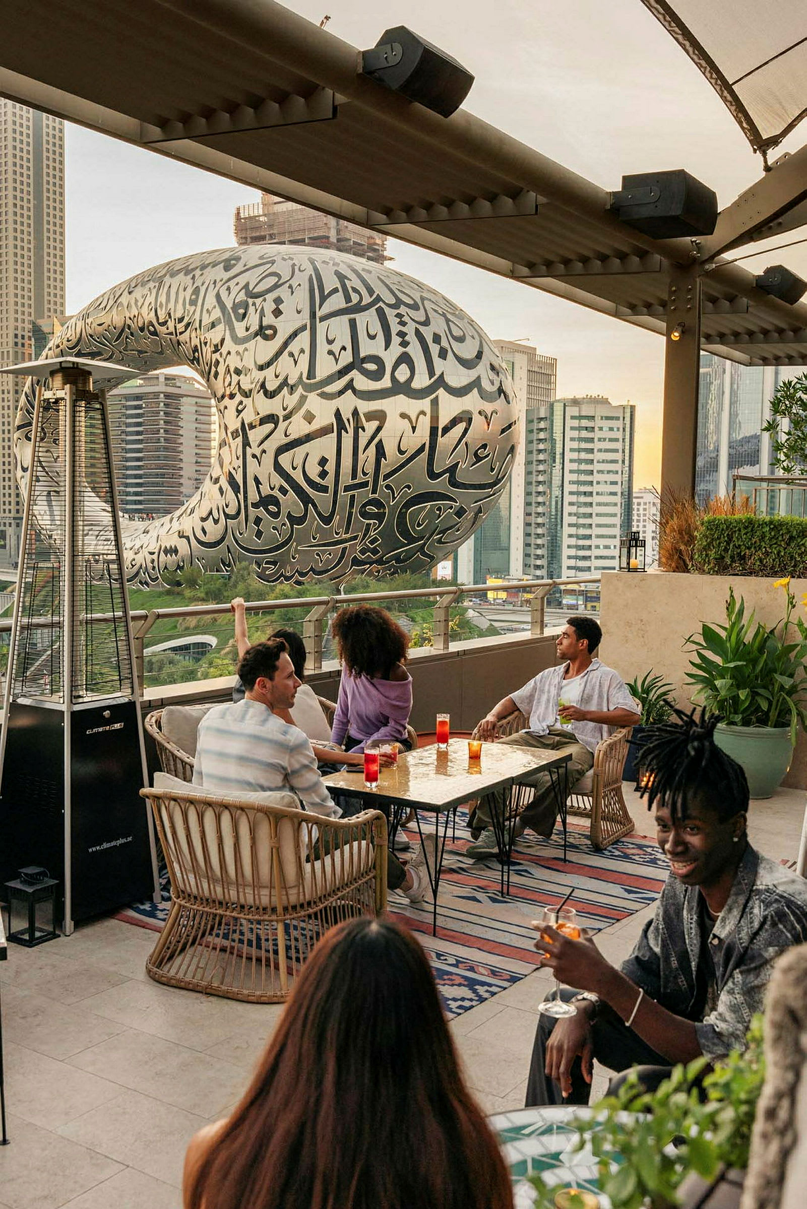 Personer, der sidder ved et bord på en tagterrasse med Museum of the Future i baggrunden. (AI-genereret tekst)
