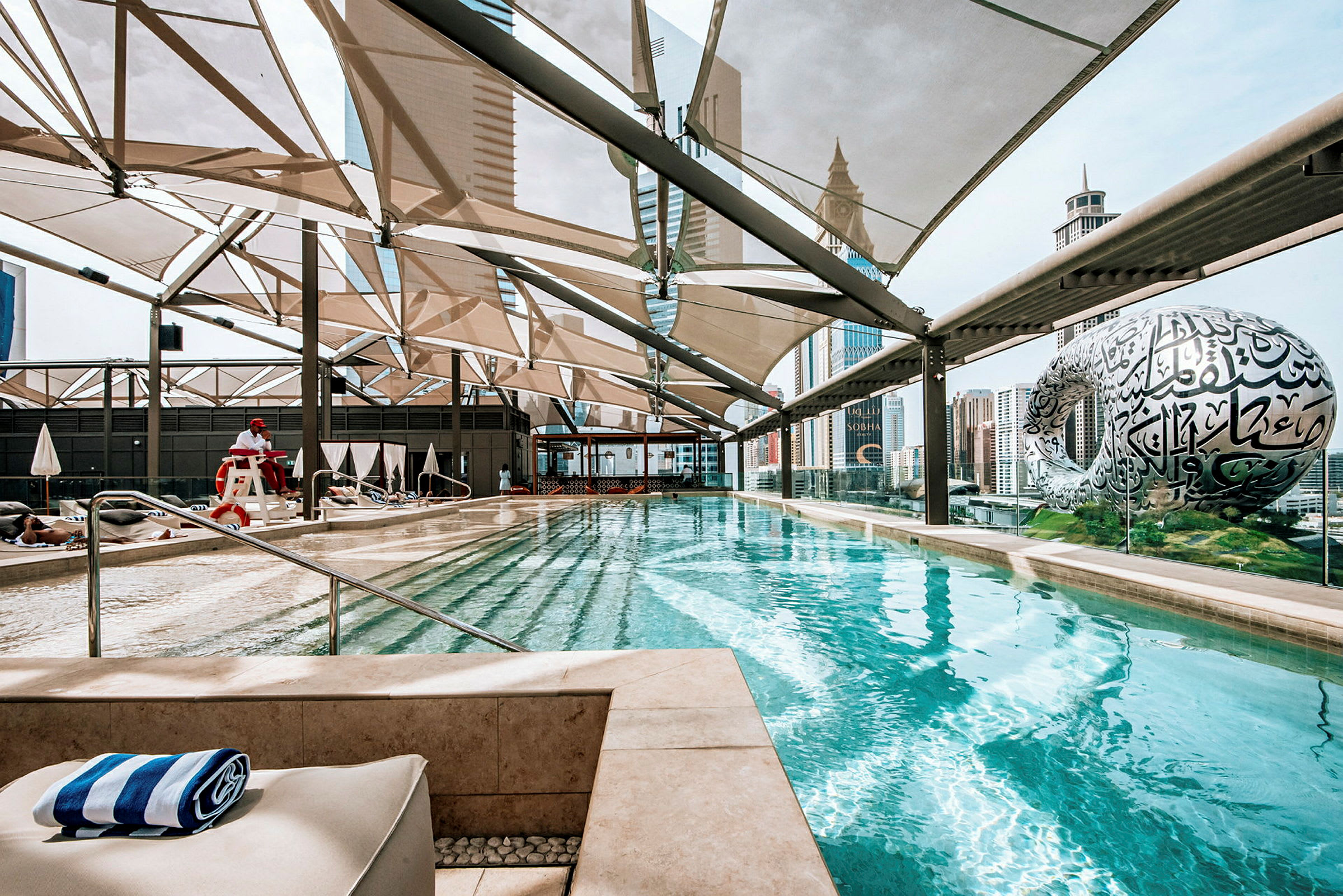 Udendørs swimmingpool med udsigt over byens skyline og en futuristisk museumsbygning. (AI-genereret tekst)