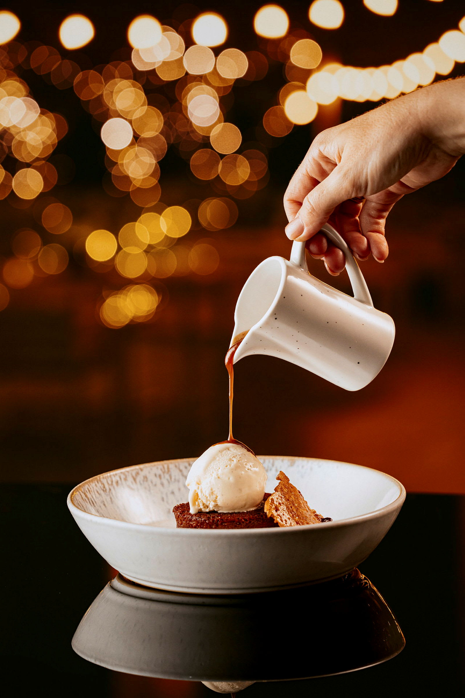 En hånd hælder sauce fra en lille kande over en dessert med is og kage i en skål, med sløret belysning i baggrunden. (AI-genereret tekst)