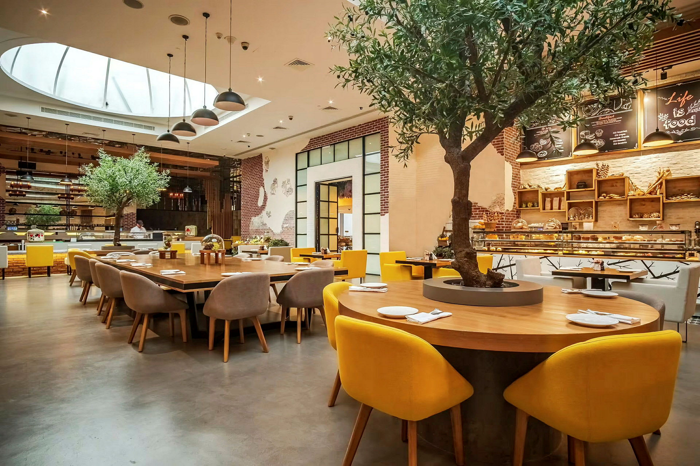 Indretning af en restaurant med et stort rundt bord omgivet af stole med gule ryglæn. Et træ er plantet midt i bordet, og baggrunden viser en serveringsdisk og dekorationer. (AI-genereret tekst)
