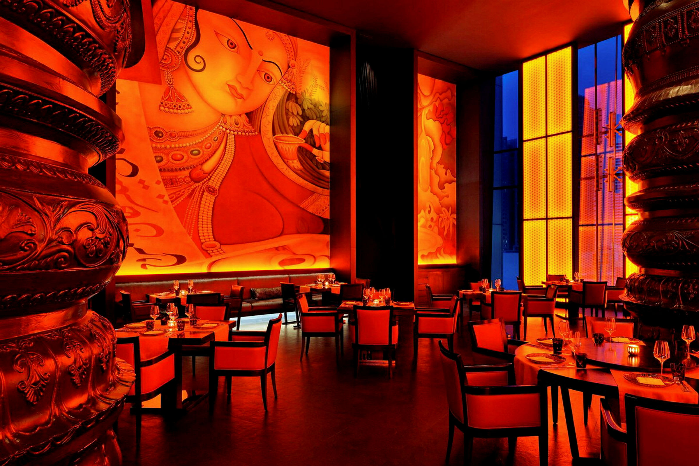 En restaurant med røde og orange farvetemaer, høje vægge med kunst og store vinduer. Møblerne består af borde og stole. (AI-genereret tekst)