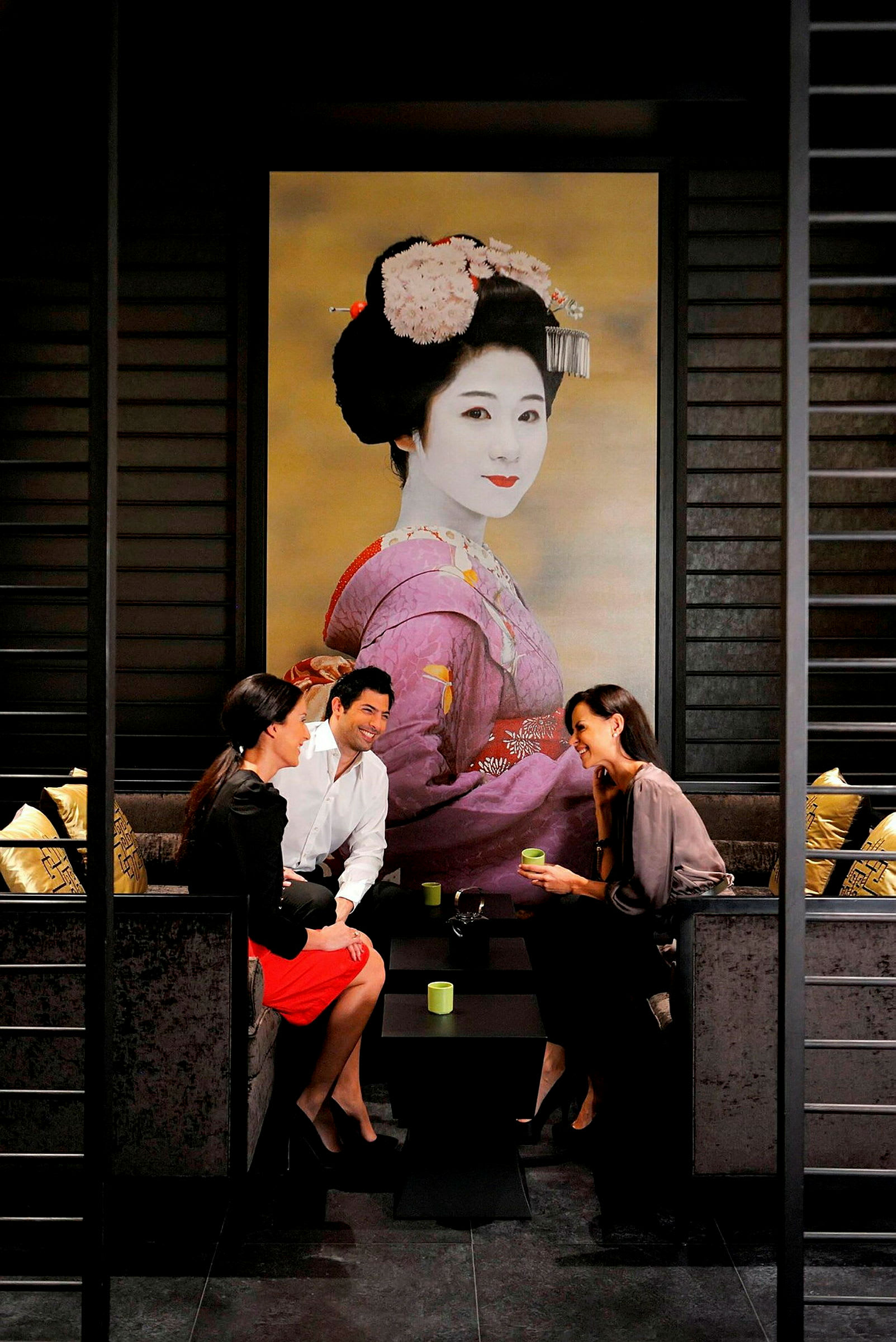 Tre personer sidder ved et bord i en elegant indretning med et stort billede af en geisha i baggrunden. (AI-genereret tekst)