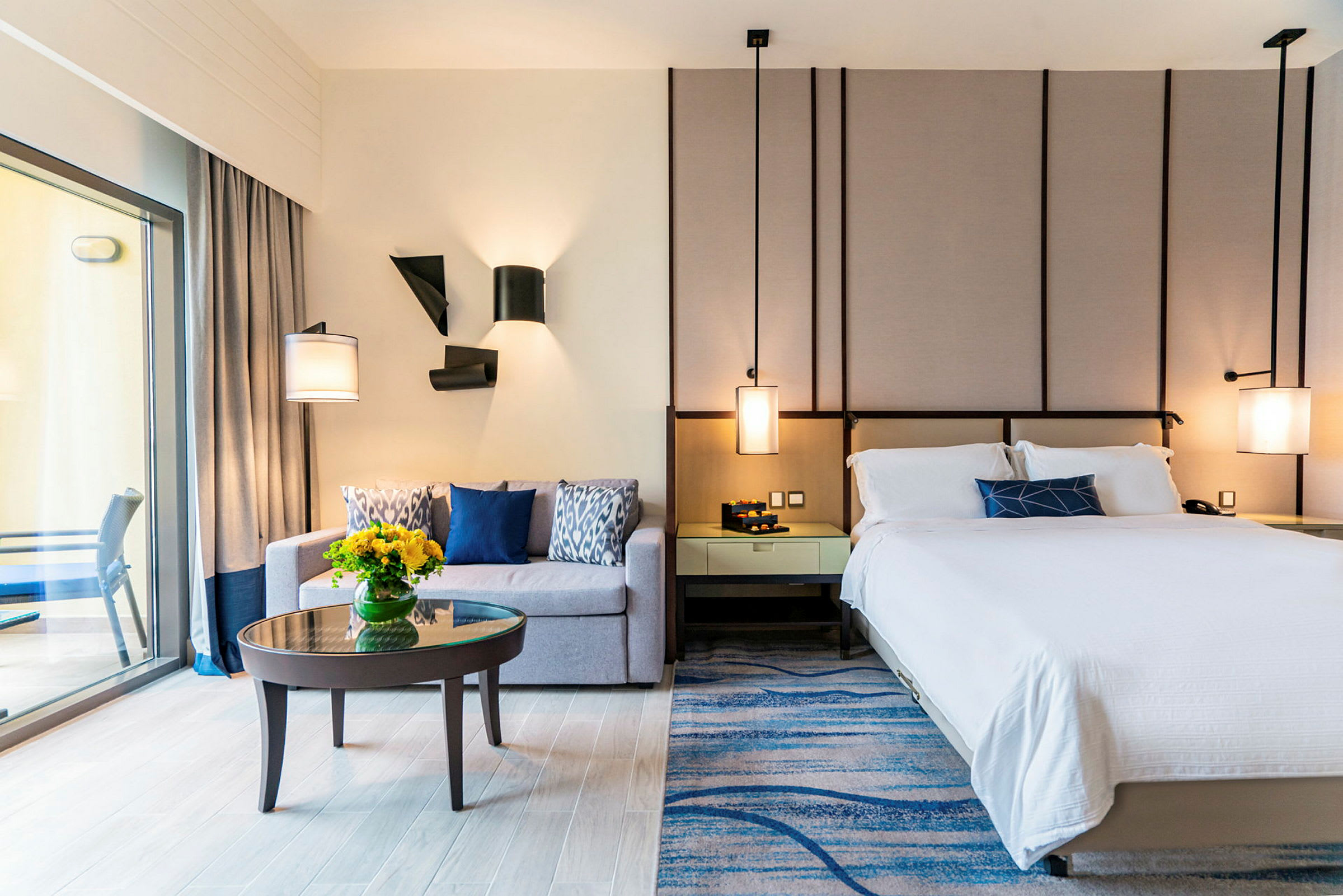 Et moderne hotelværelse med en seng, en sofa, et lille sofabord med en vase med blomster og en altan, der er synlig gennem en glasdør. (AI-genereret tekst)