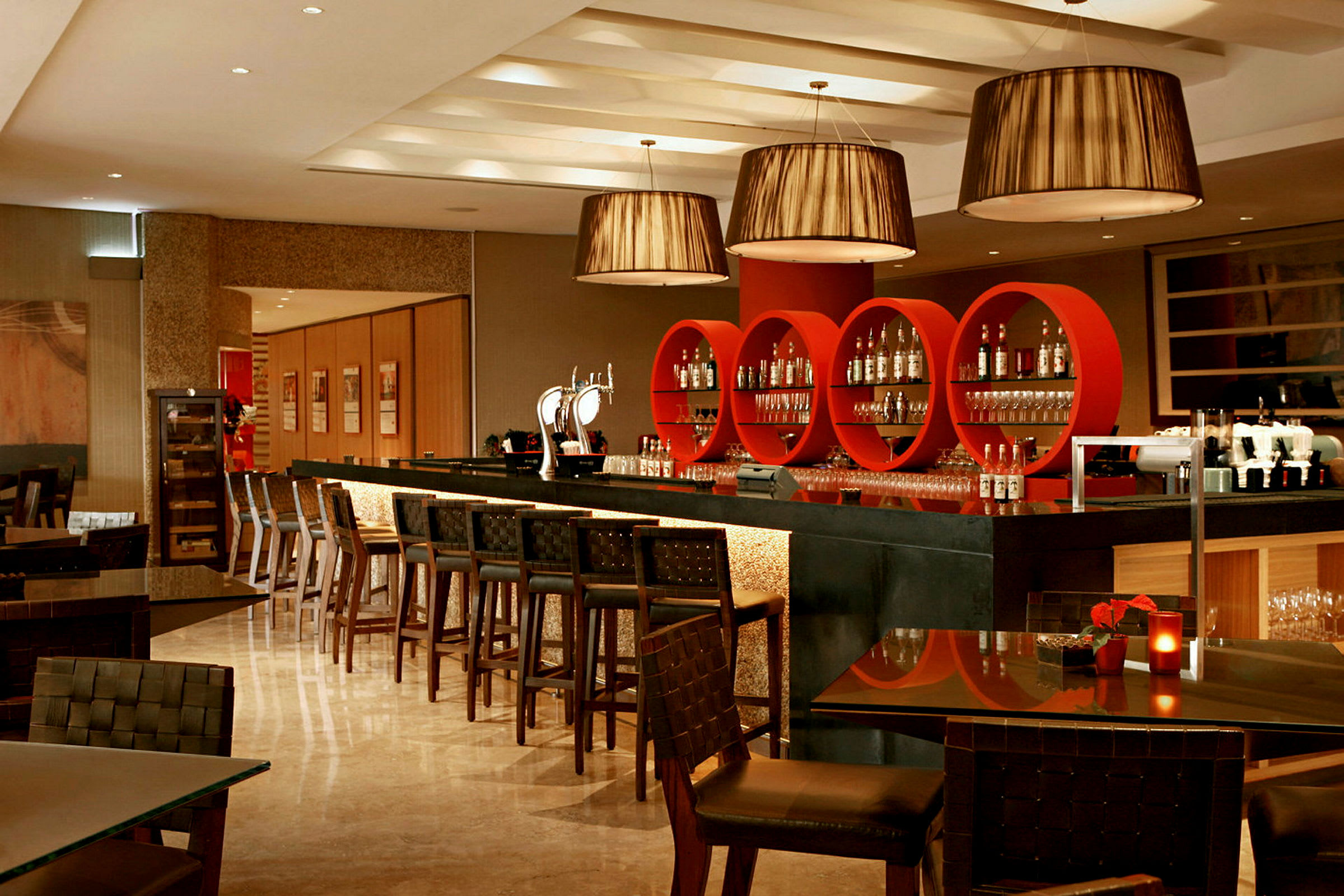 Billede af en moderne bar med røde, cirkelformede hylder og belysning. Bartenderdisk og barstole samt borde med stole i forgrunden. (AI-genereret tekst)