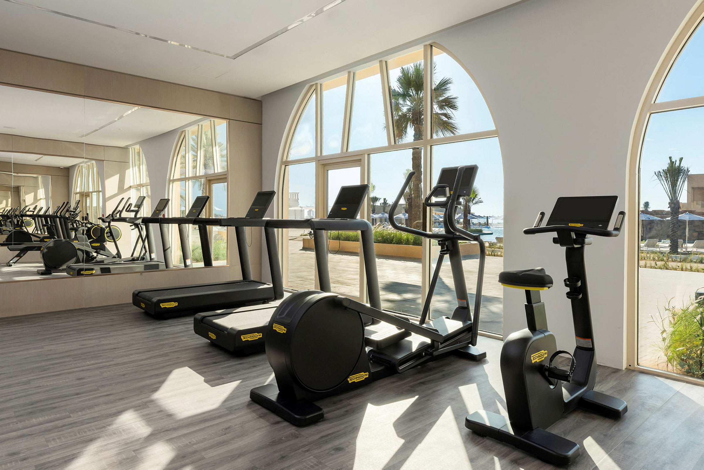 Træningsmaskiner i et fitnesscenter med vinduer ud mod udendørsområdet. (AI-genereret tekst)