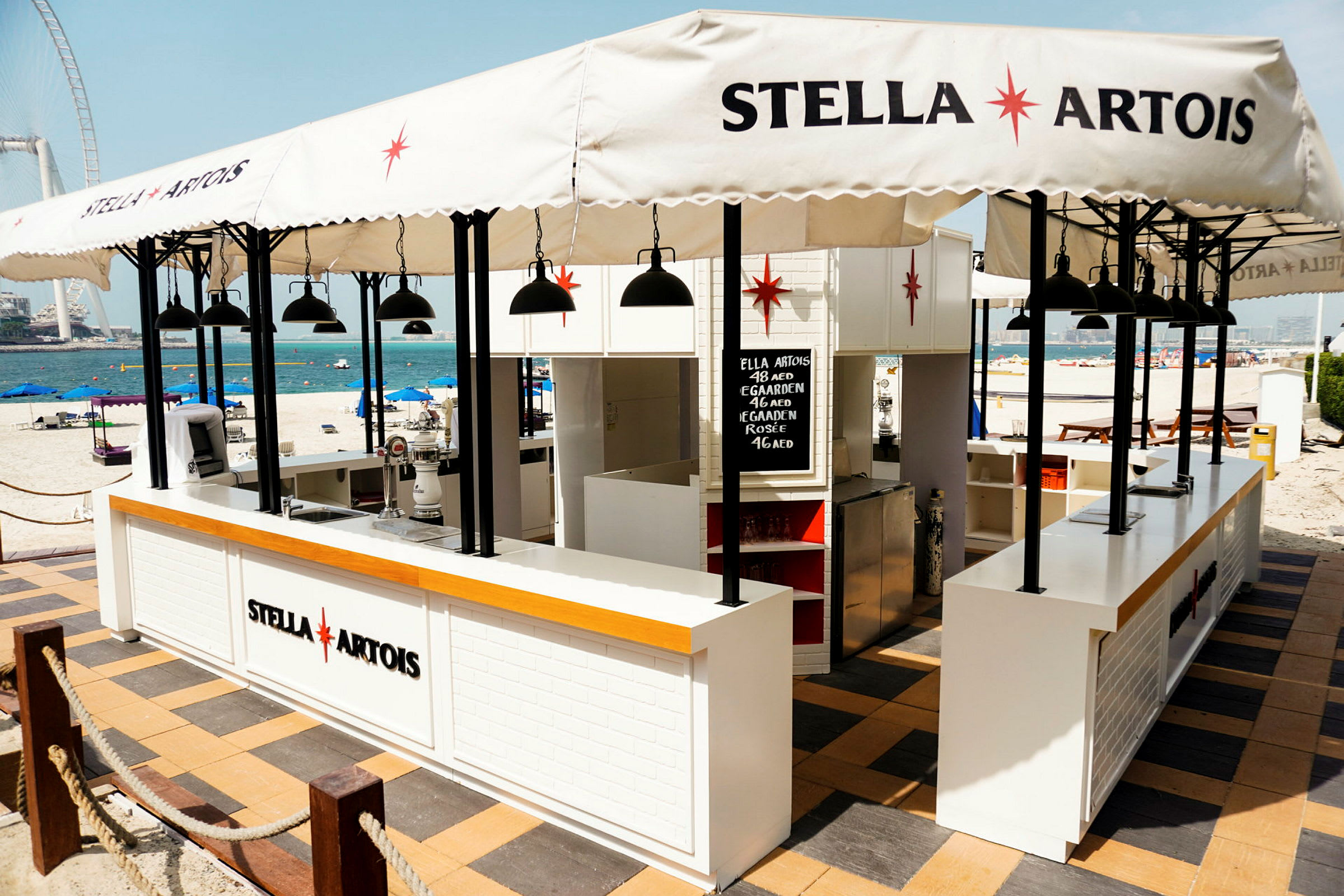 En strandbar med Stella Artois-logo, parasoller og borde. (AI-genereret tekst)
