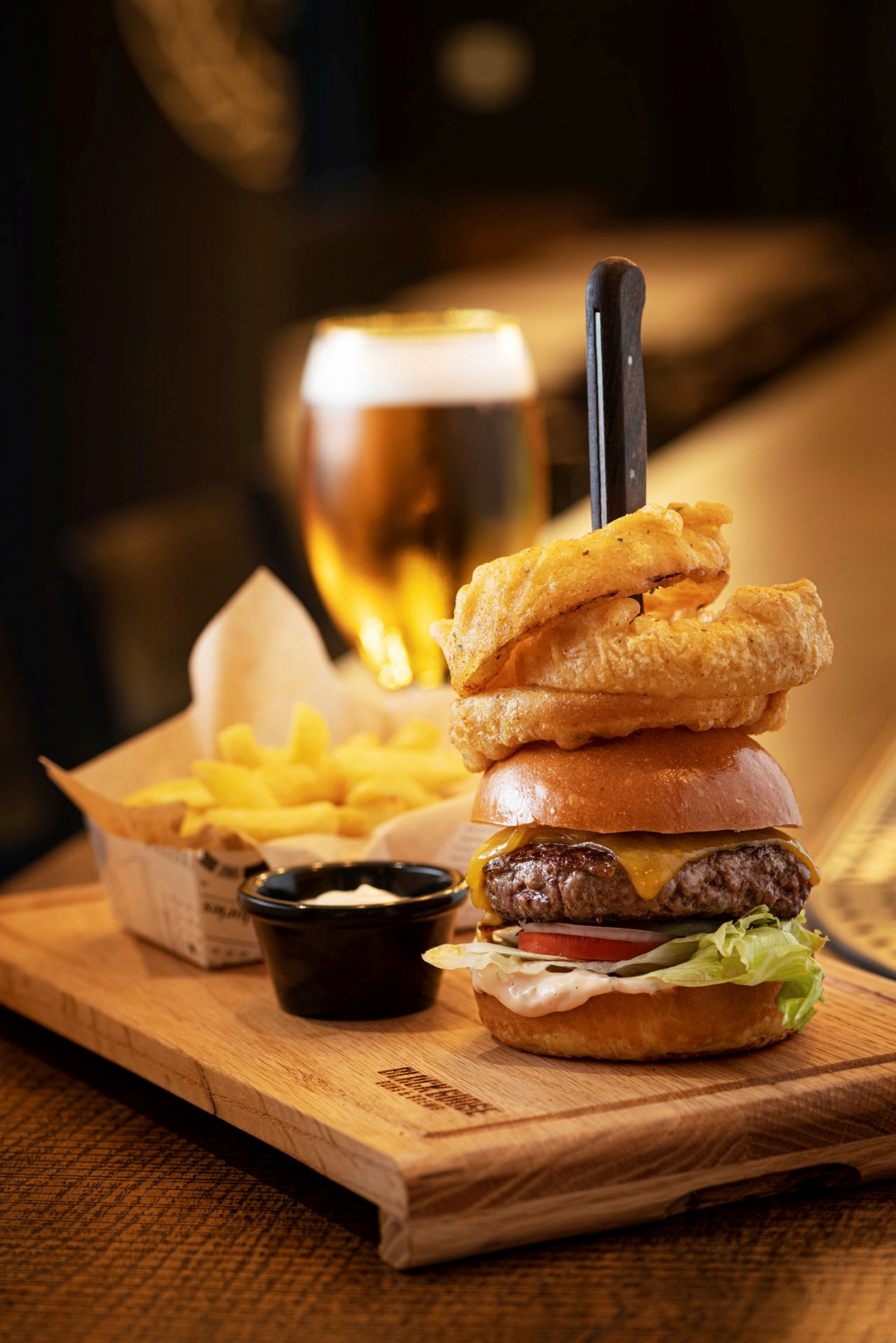 En tallerken med en burger, løgringe, pommes frites og en dip. Et glas øl ses i baggrunden. (AI-genereret tekst)