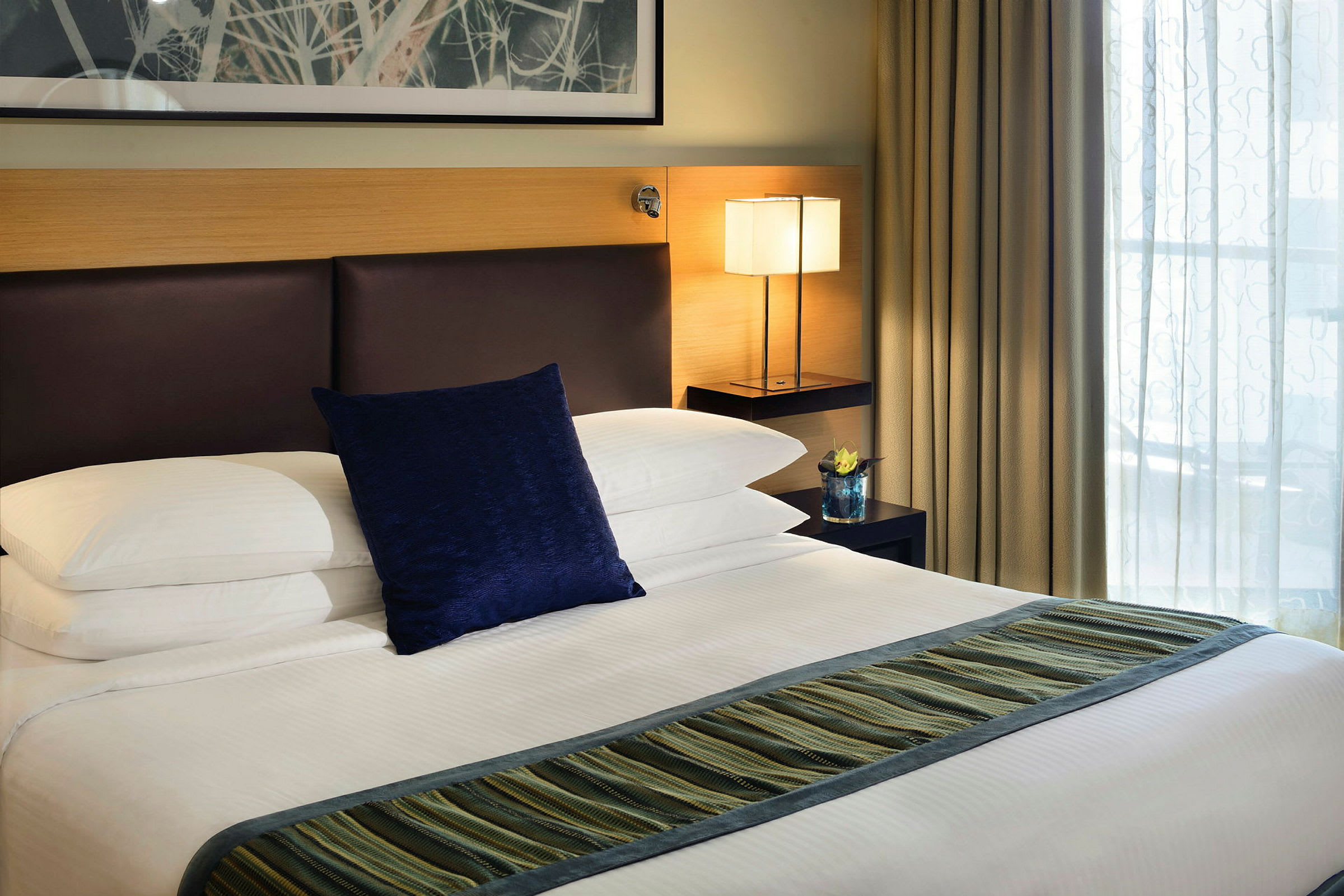 En hotelseng med hvide lagner, en blå pude og en stribet løber. Et natbord med en lampe og en dekoration er placeret ved siden af. (AI-genereret tekst)