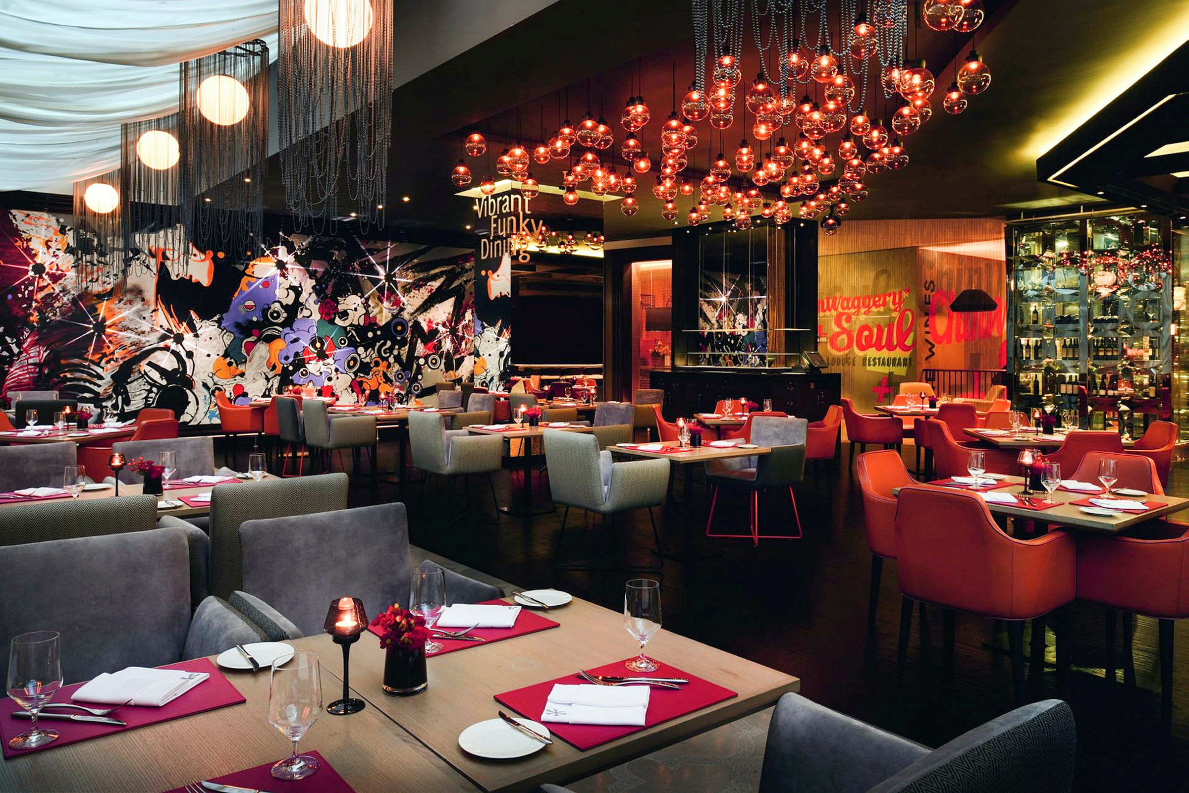 Restaurant med farverigt og moderne interiør, borde med røde servietter og glas samt lamper hængende fra loftet. (AI-genereret tekst)
