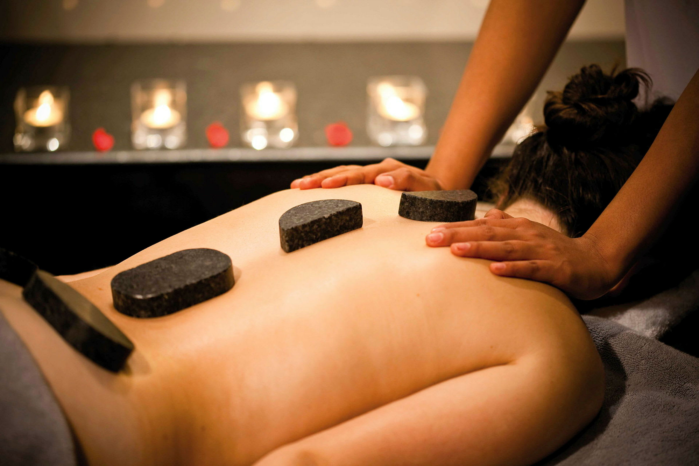 En person modtager hot-stone massage, med varme sten placeret på ryggen og hænder der giver massage. (AI-genereret tekst)