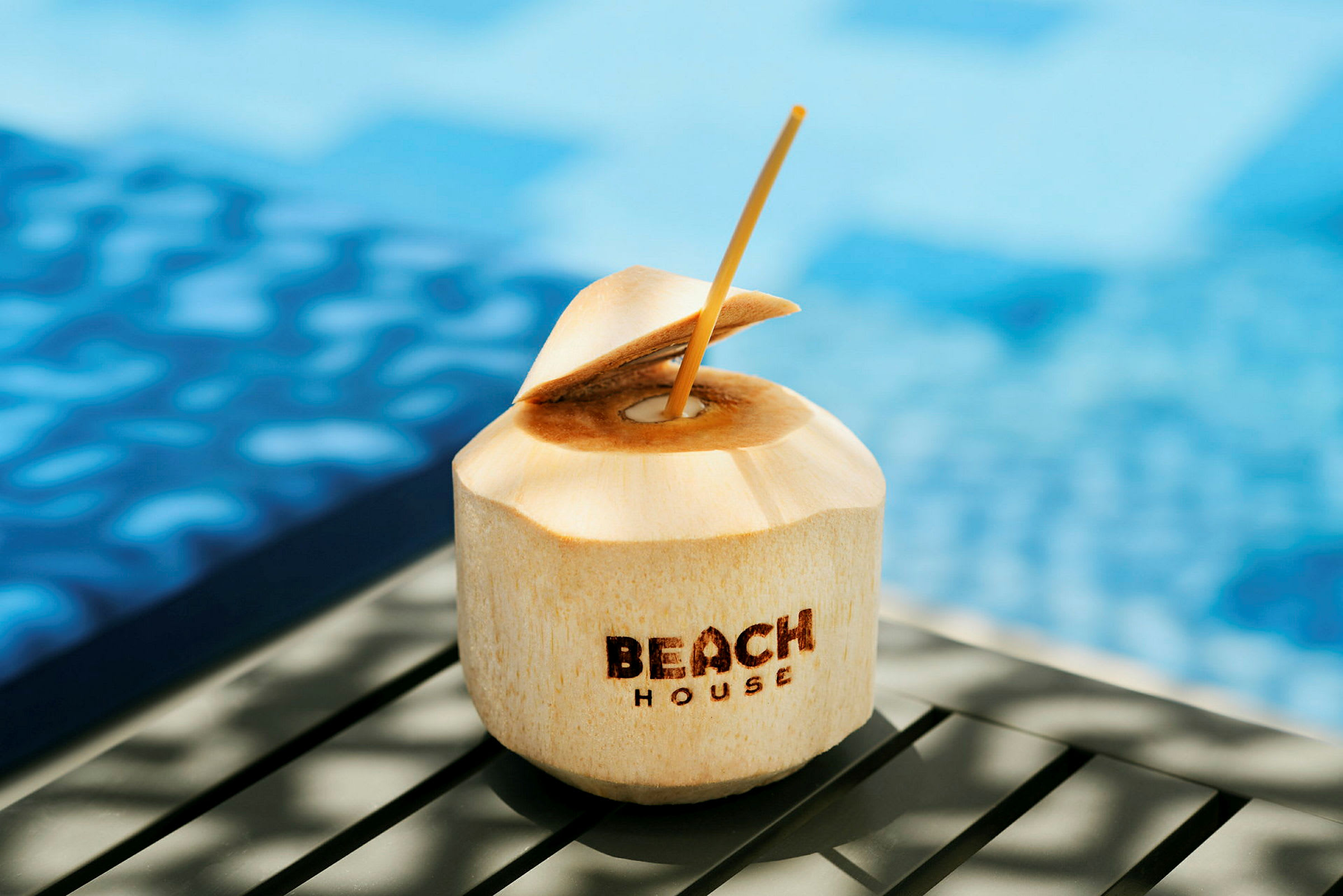En kokosnød med teksten 'Beach House' placeret nær en pool. (AI-genereret tekst)
