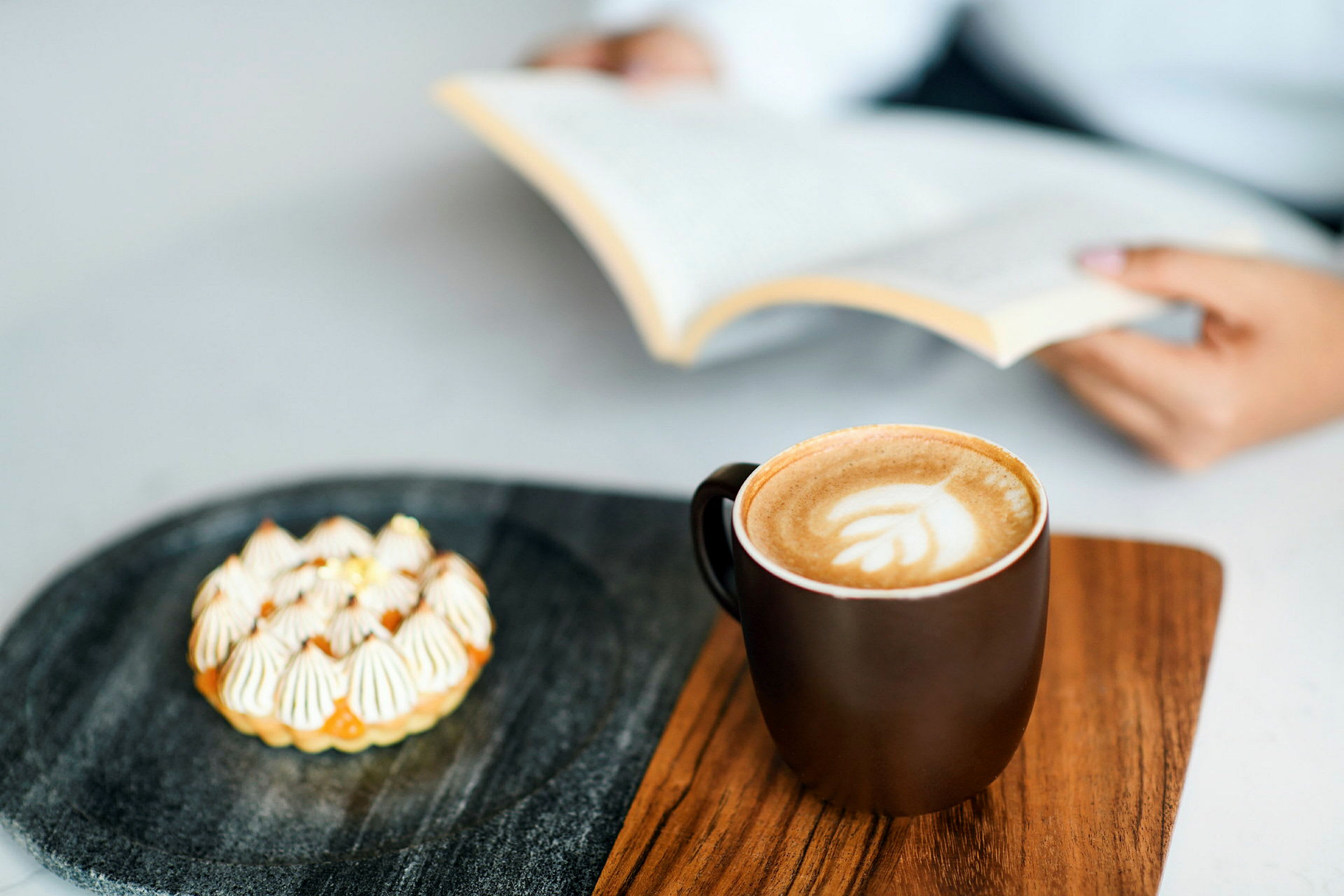 En kop kaffe og en lille kage ligger på en træbakke. En person holder en åben bog i baggrunden. (AI-genereret tekst)