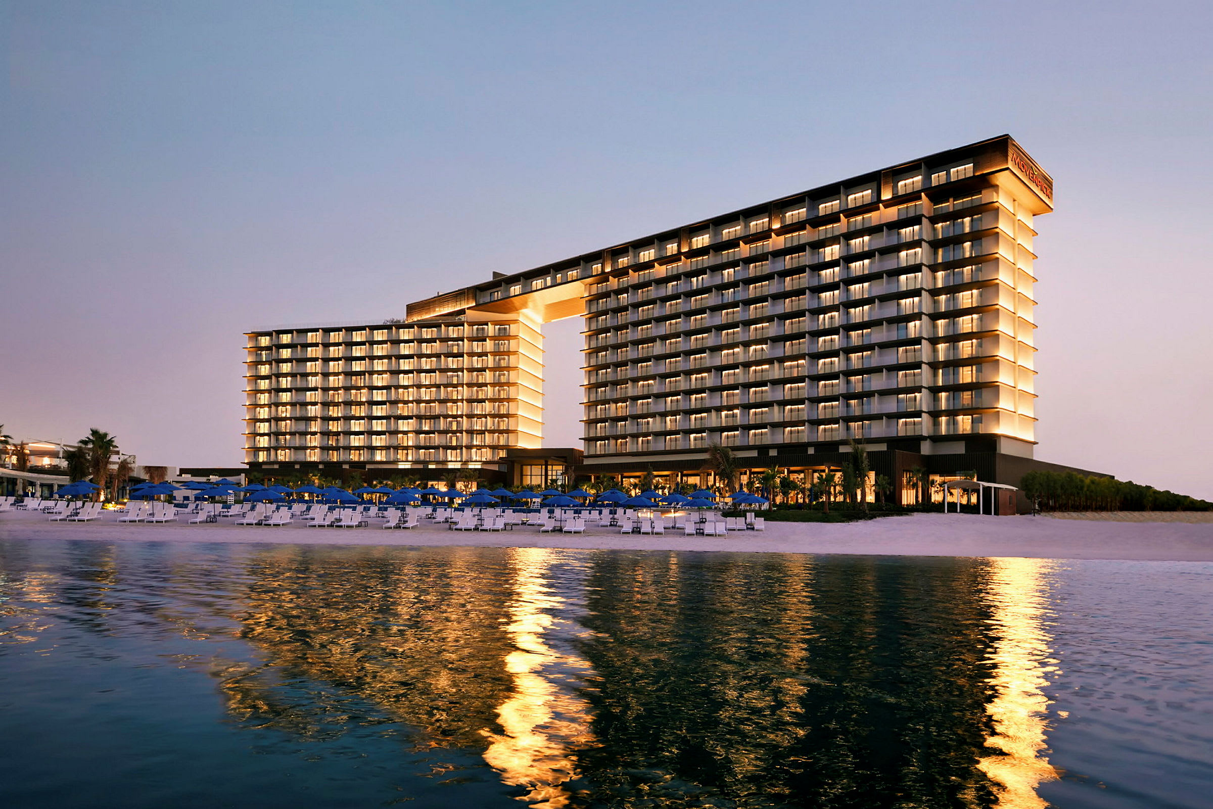 Bygningen er et moderne hotelkompleks ved en strand, oplyst og reflekterer sig i vandet. (AI-genereret tekst)