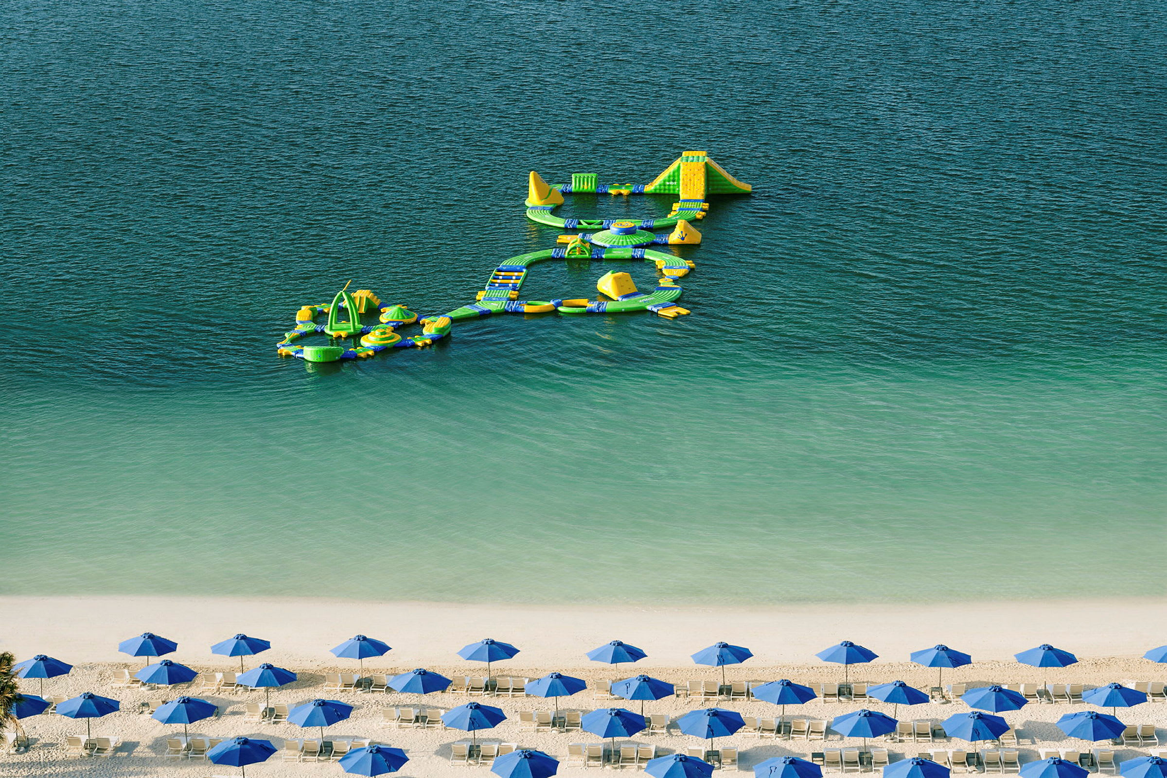 En strand med hvide liggestole og blå parasoller foran klart vand, hvor en oppustelig forhindringsbane flyder. (AI-genereret tekst)