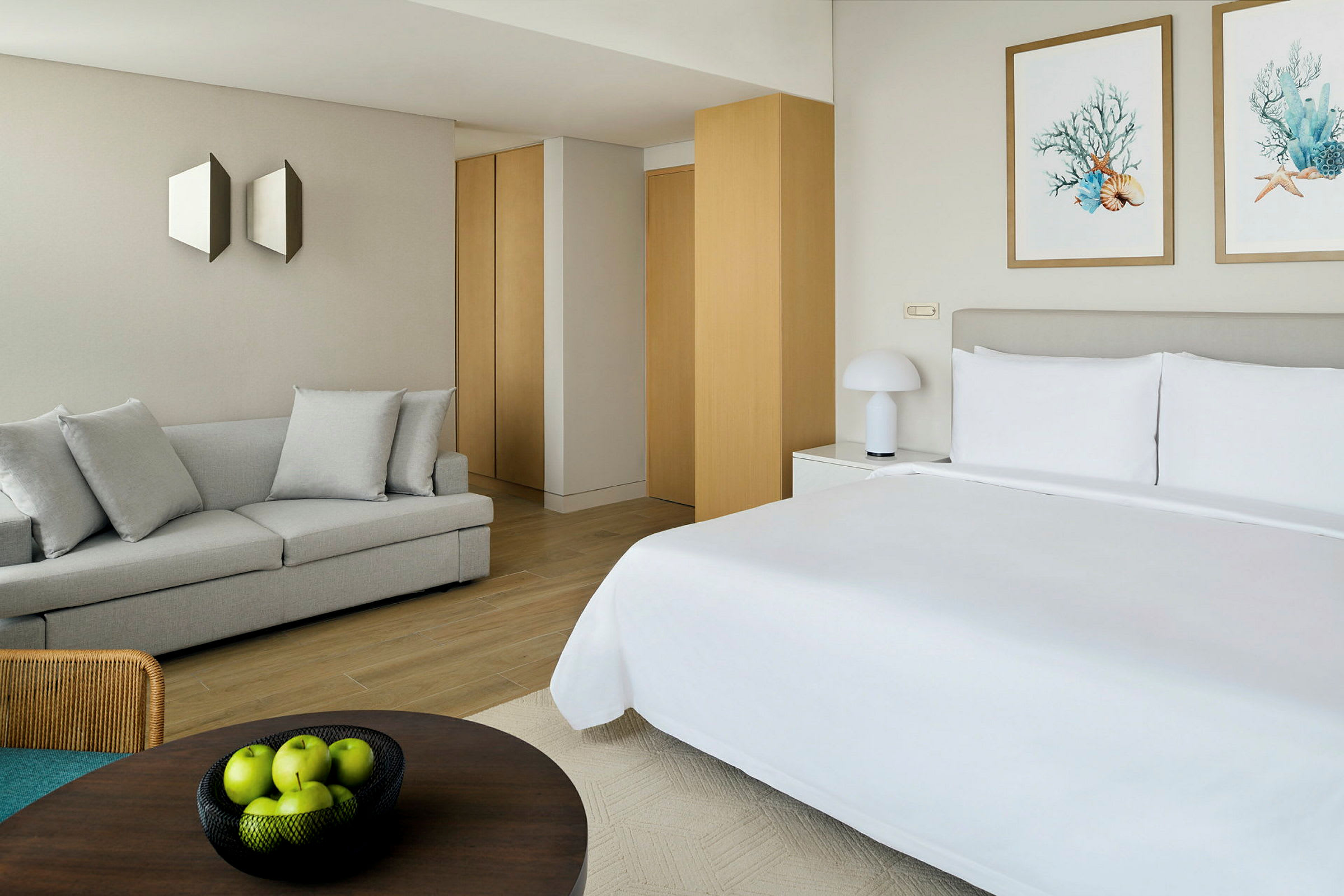 Et moderne hotelværelse med en opredt dobbeltseng. Værelset indeholder også en lys sofa, vægdekorationer og en skål med grønne æbler på et bord. (AI-genereret tekst)