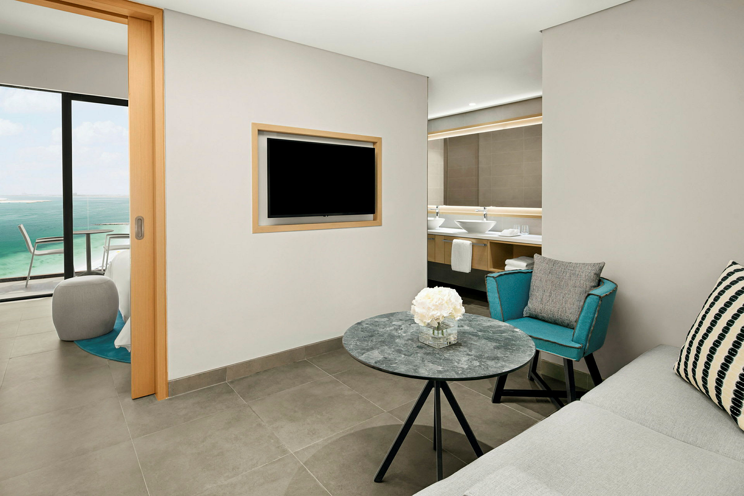 Et hotelværelse med siddeområde, et bord med blomster, en sofa, en lænestol og et TV monteret på væggen. I baggrunden ses et badeværelse og gennem en glasskydedør en udsigt over havet. (AI-genereret tekst)