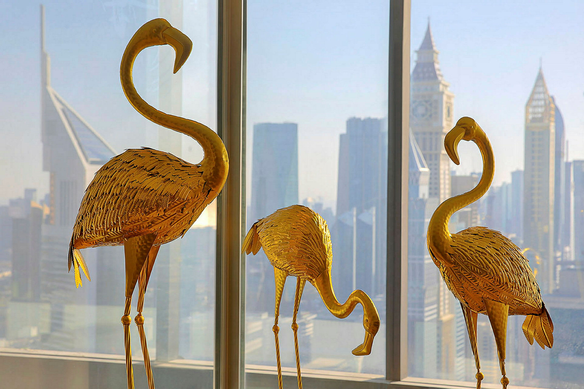 Tre guldfarvede flamingoskulpturer foran et vindue med en by-skyline i baggrunden. (AI-genereret tekst)