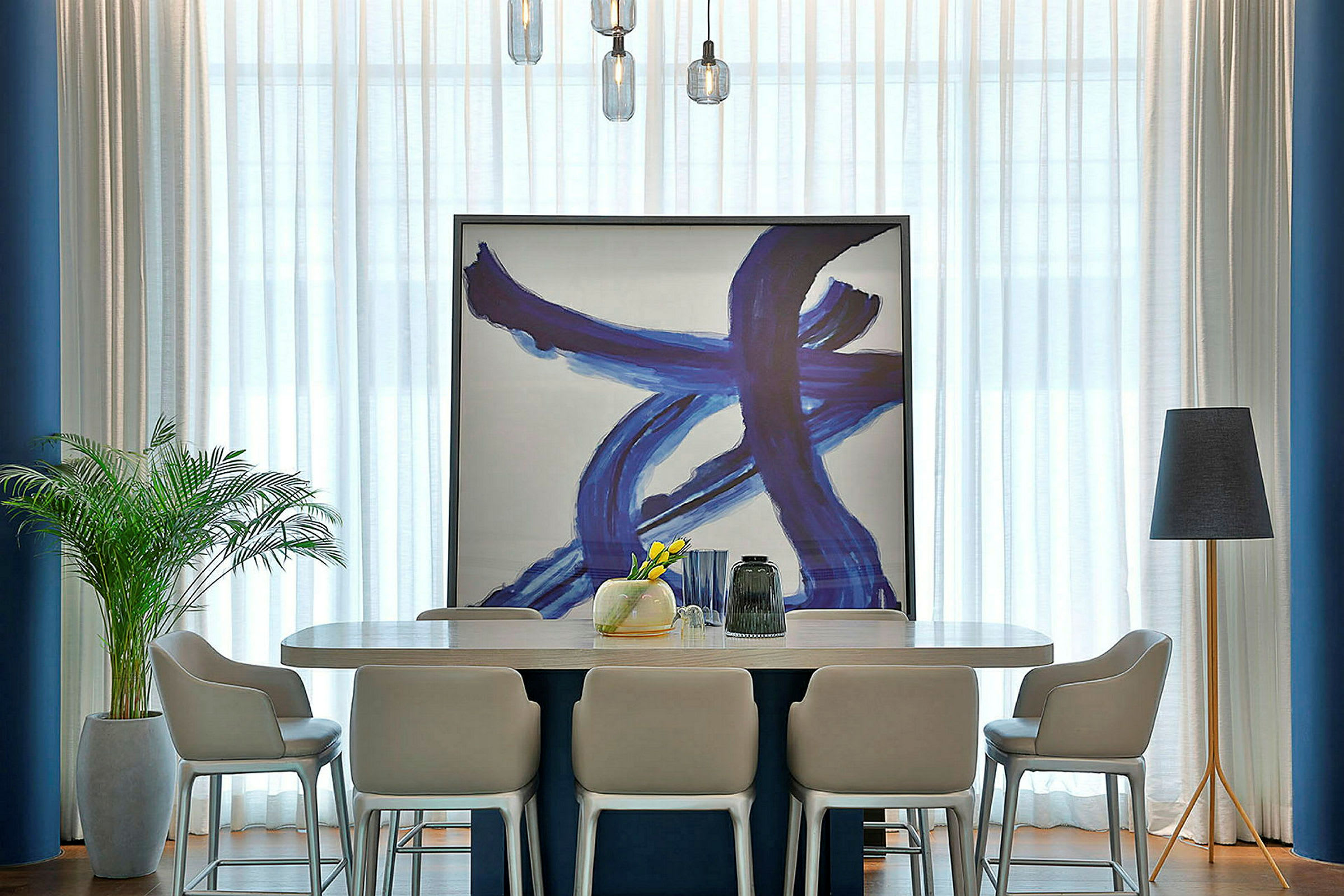 Interiør med et moderne spisebord omgivet af stole. Et stort kunstværk med blå penselstrøg er opstillet mod væggen. En gulvlampe og en indendørsplante er placeret på hver side af bordet. (AI-genereret tekst)