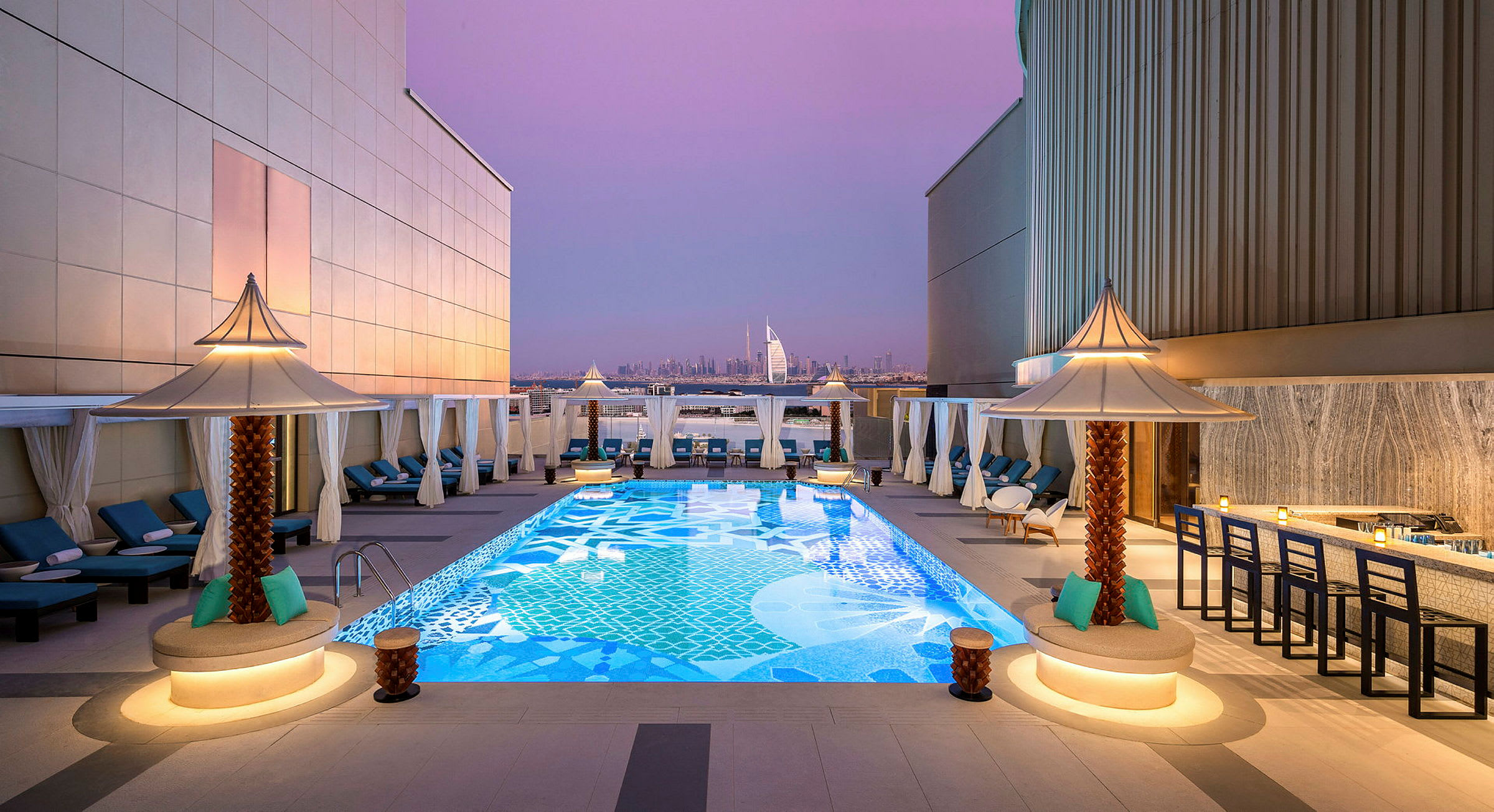 En tagterrasse med en dekorativ pool omgivet af liggestole og små parasoller. I baggrunden kan Burj Al Arab og Dubais skyline ses under en lilla aftenshimmel. (AI-genereret tekst)