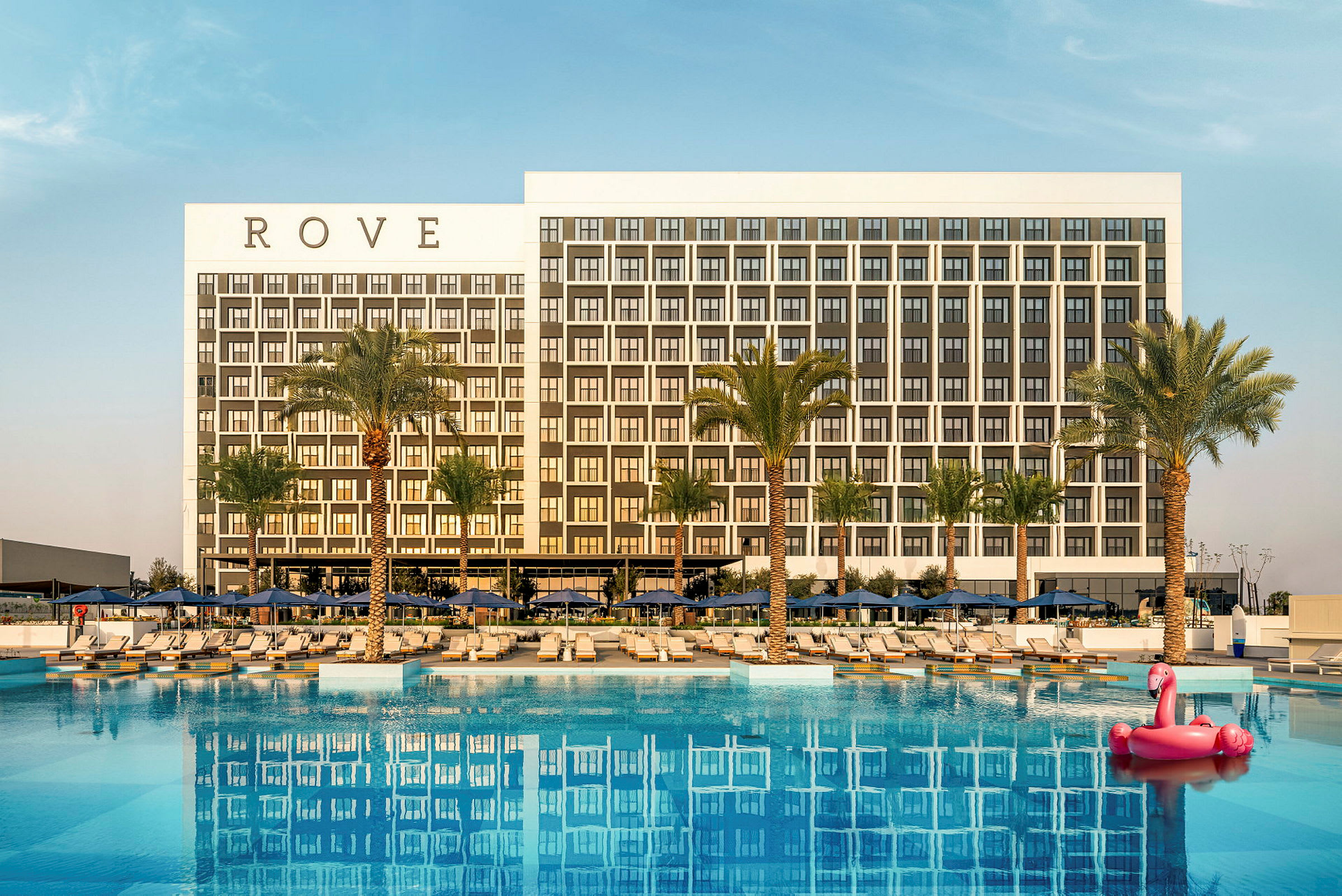 Billede af et moderne hotel med en stor pool og palmer i forgrunden. En lyserød flamingo på en flydende legetøj ses i poolen. (AI-genereret tekst)