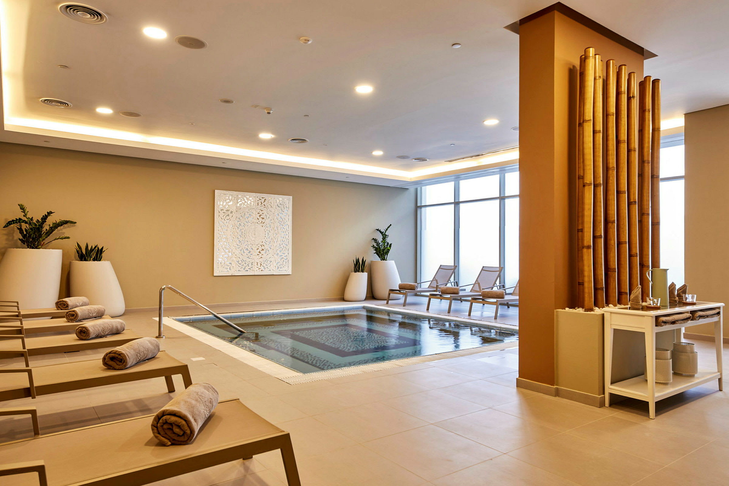 Et indendørs spa-område med en pool, liggestole med håndklæder og dekorative bambusstænger. (AI-genereret tekst)