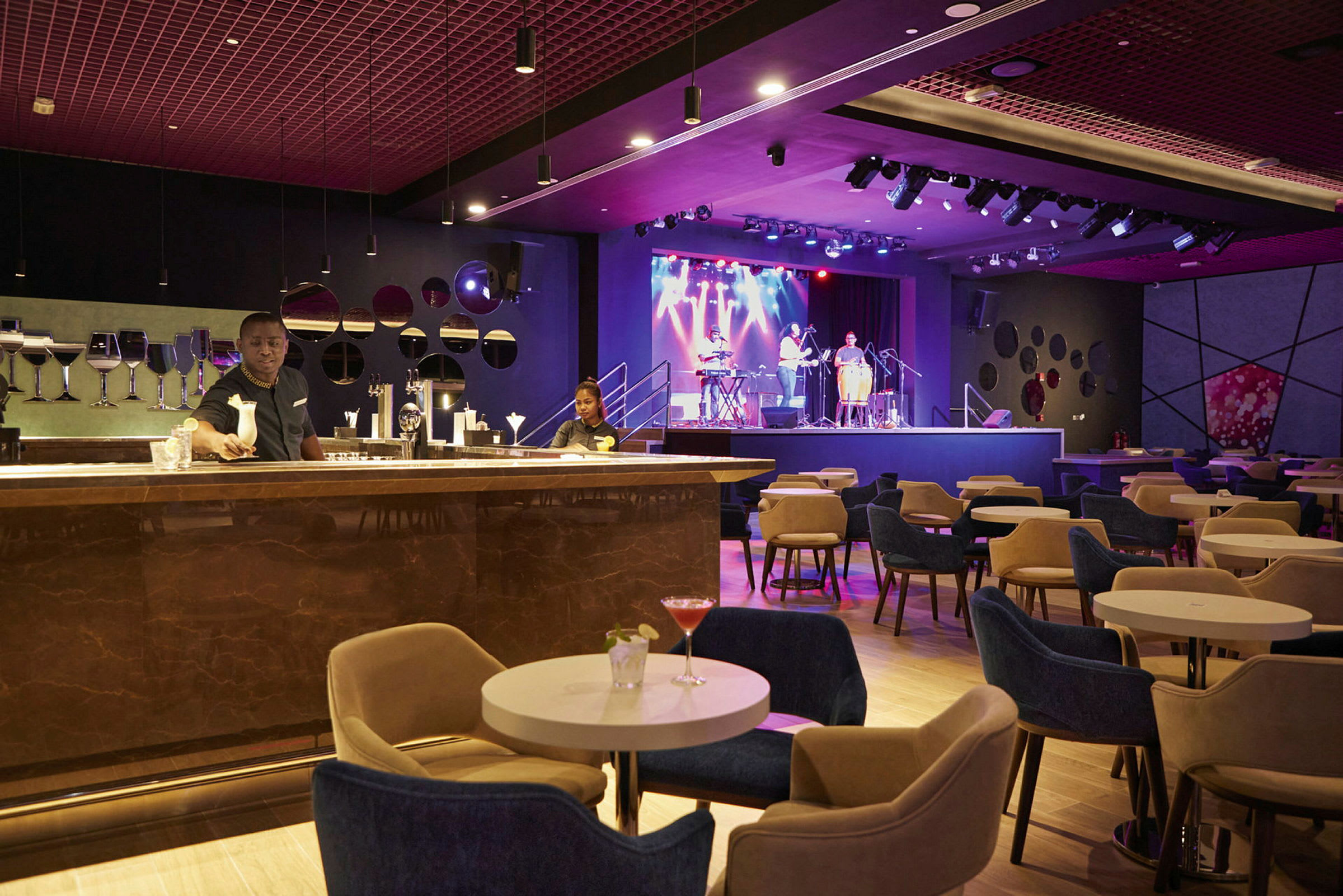 En elegant lounge med siddepladser omkring små borde, en bardisk i forgrunden og en scene med musikere i baggrunden. (AI-genereret tekst)