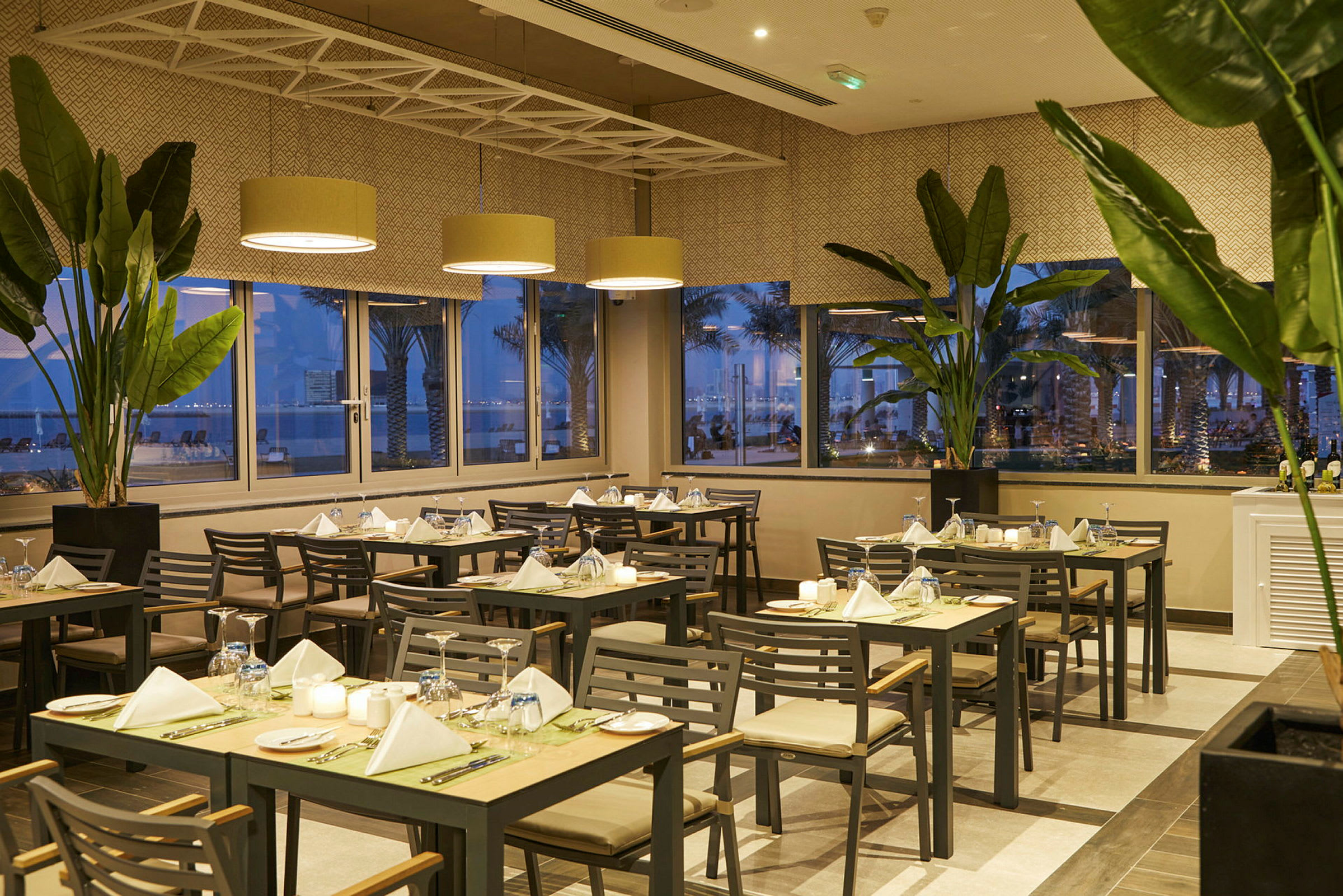 Restaurantmiljø med dækkede borde, grønne planter og panoramavinduer med aftenudsigt. (AI-genereret tekst)