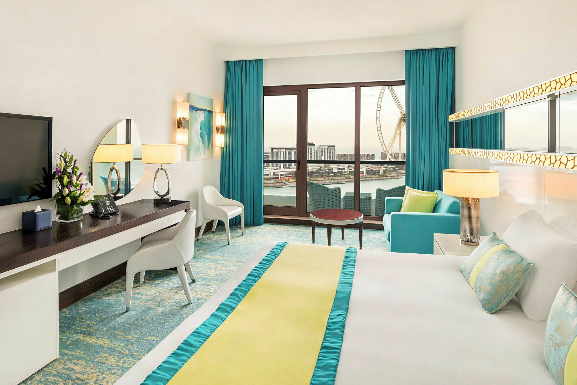 Moderne hotelværelse med lyse farver, stor seng, skrivebord og siddeområde. Et stort vindue med udsigt over en stor by og et pariserhjul. (AI-genereret tekst)