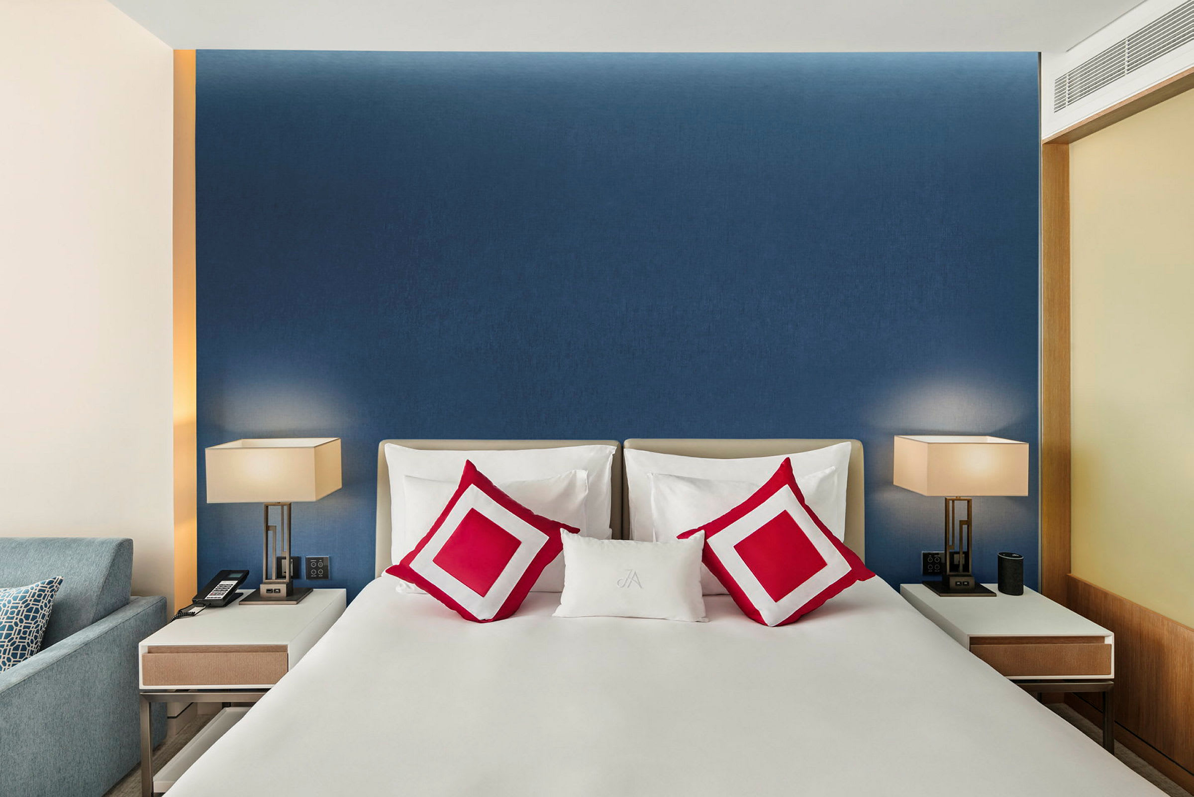 Et moderne, stilfuldt hotelværelse med en dobbeltseng med hvidt sengetøj og røde pyntepuder. På hver side af sengen er der hvide, elegante natborde med bordlamper. (AI-genereret tekst)