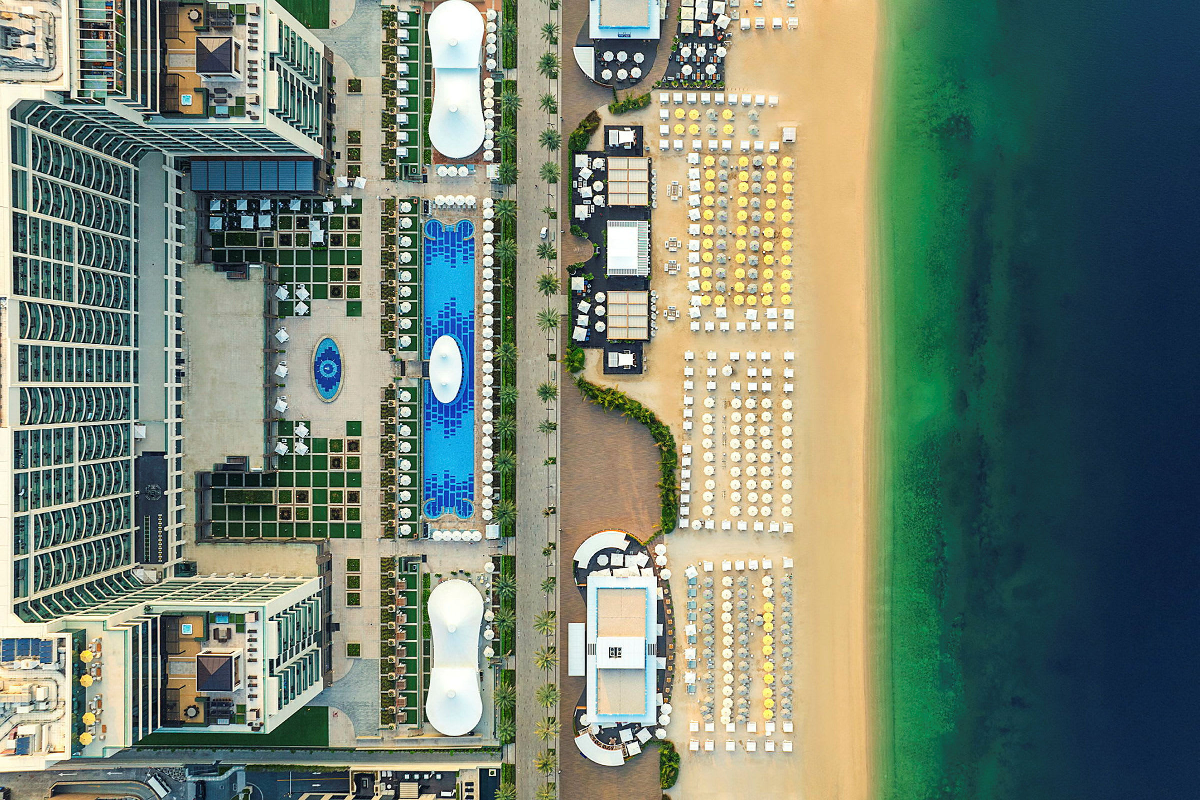 Luftfoto af hotelområde med pools og liggestole ved siden af strand og hav. (AI-genereret tekst)