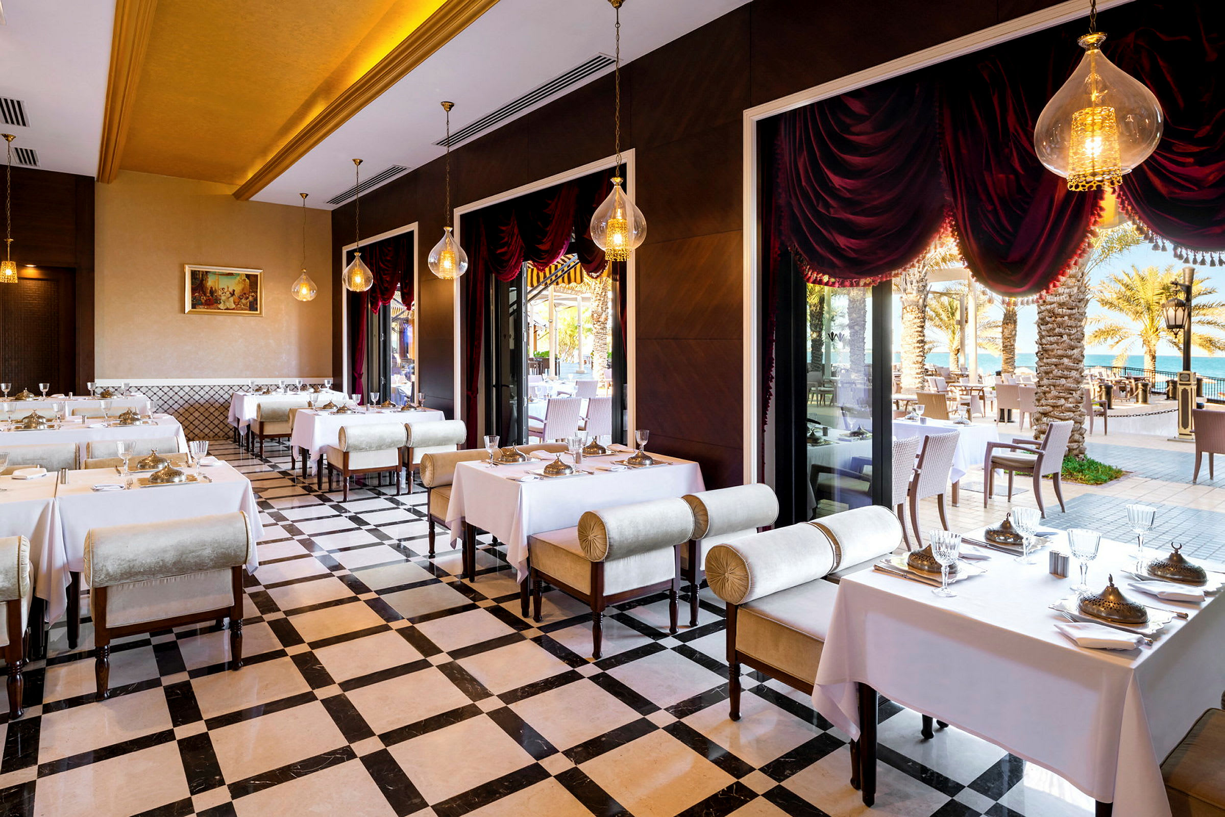 En elegant restaurant med dækkede borde, store vinduer og udsigt til palmer og hav. (AI-genereret tekst)