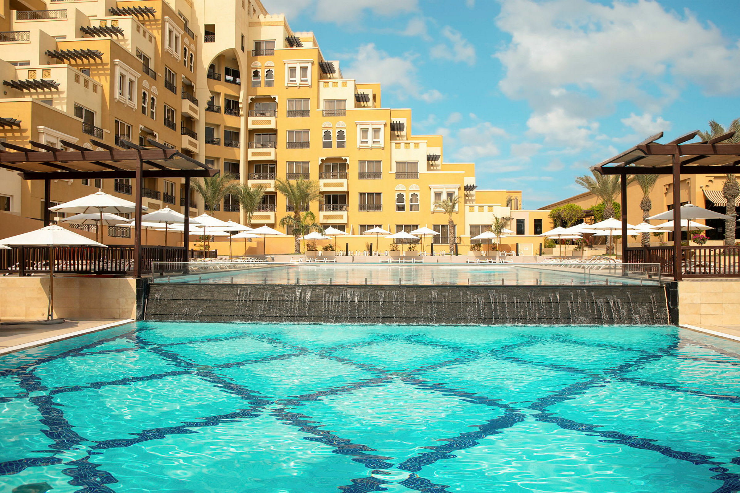 Et hotel med gul facade, pool og parasoller. (AI-genereret tekst)