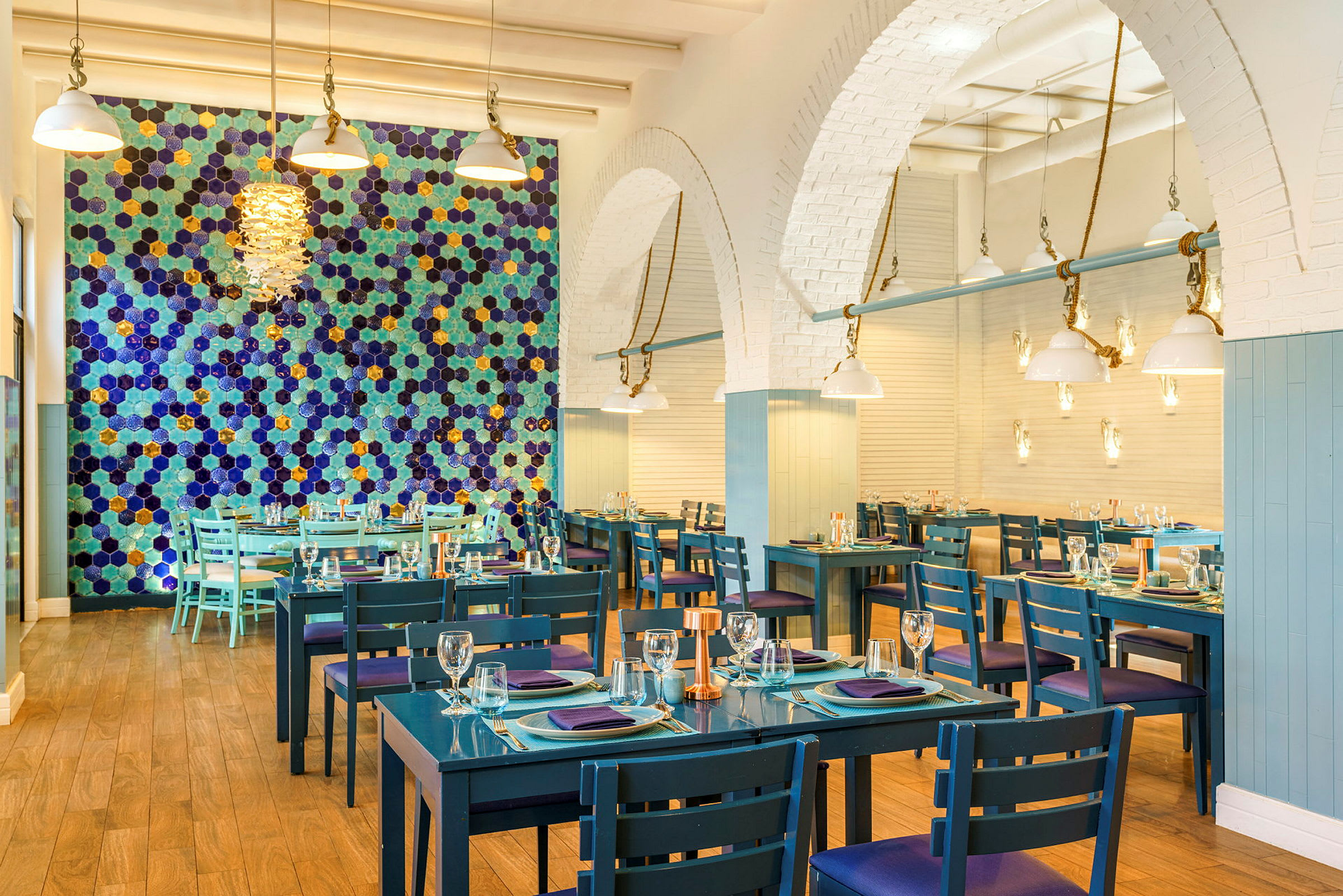 Billede af en restaurant med blå borde og stole, en væg med farverige sekskantede mønstre og moderne loftslamper. (AI-genereret tekst)