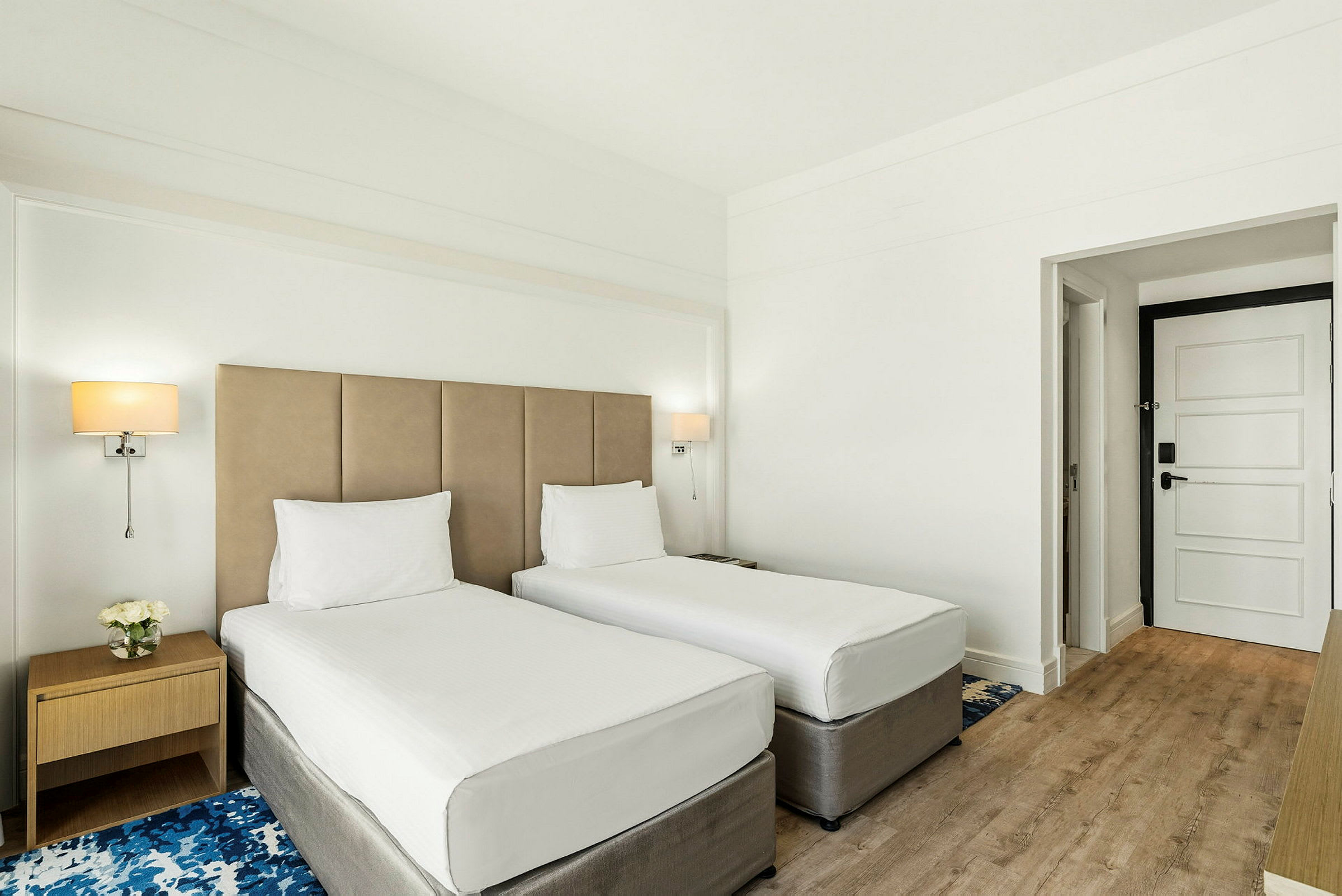 Et hotelværelse med to enkeltsenge, et natbord og en dør. (AI-genereret tekst)