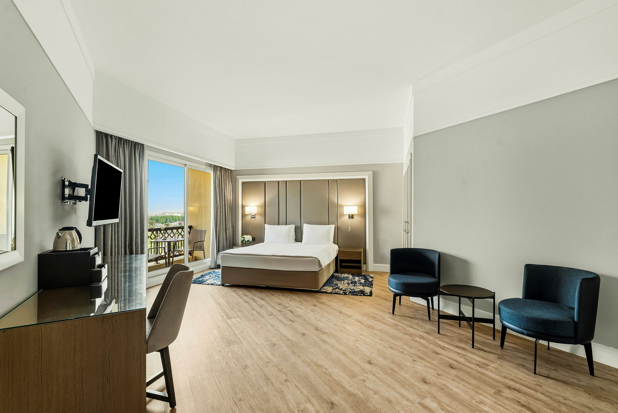 Et moderne hotelværelse med en dobbeltseng, en siddegruppe med stole og bord samt en arbejdsplads. Udgang til balkon synlig gennem store vinduer. (AI-genereret tekst)