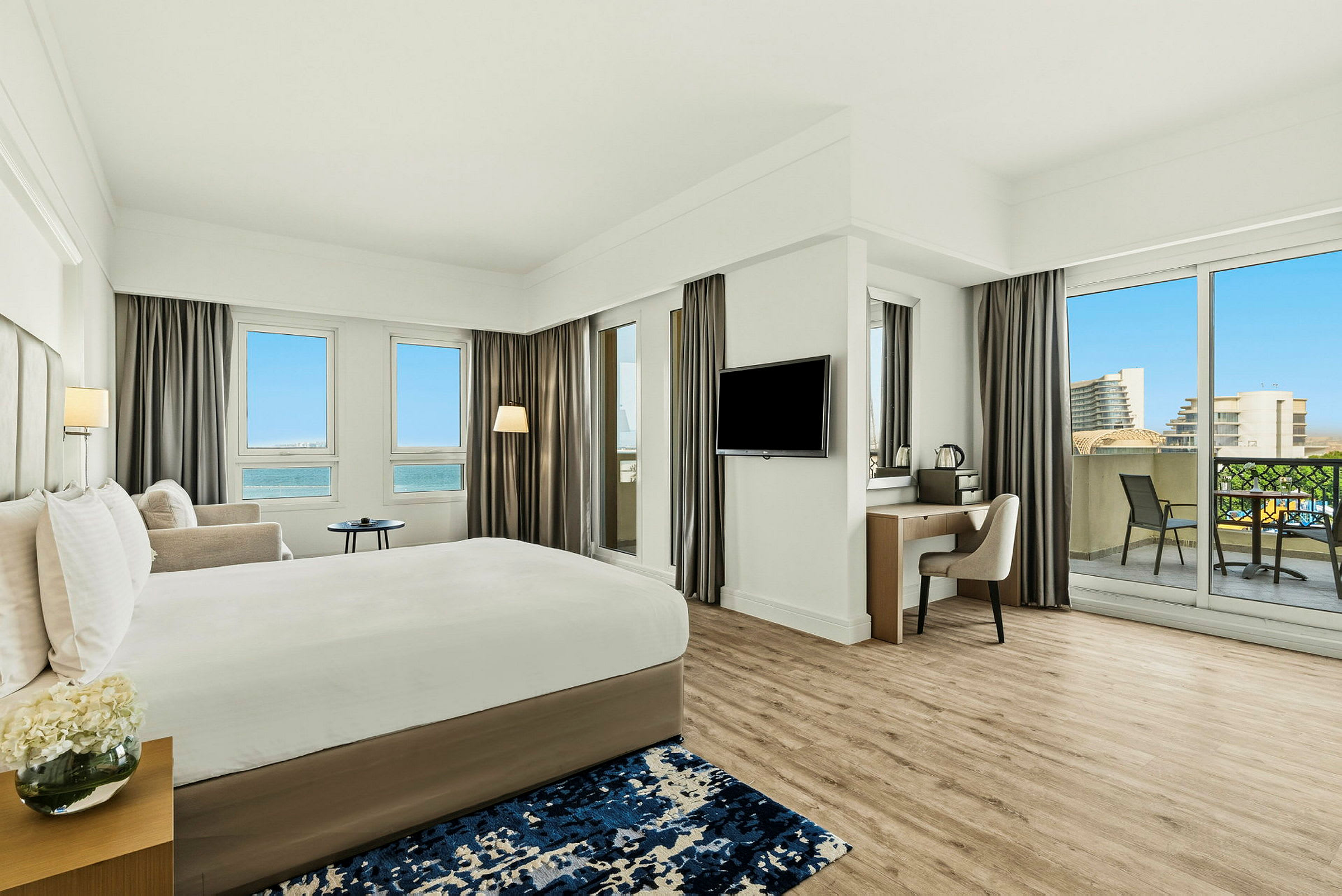 Et moderne hotelværelse med en stor seng, et skrivebord, balkon og udsigt over en blå himmel og bygninger. (AI-genereret tekst)